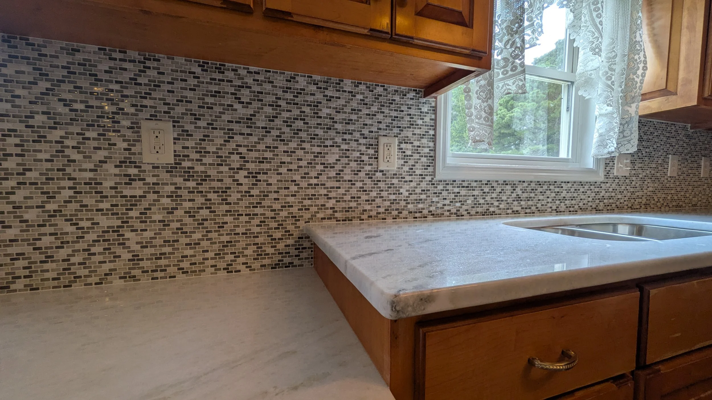 Kitchen countertop with a mosaic tile backsplash, a double electrical outlet, and a window with lace curtains above the sink.