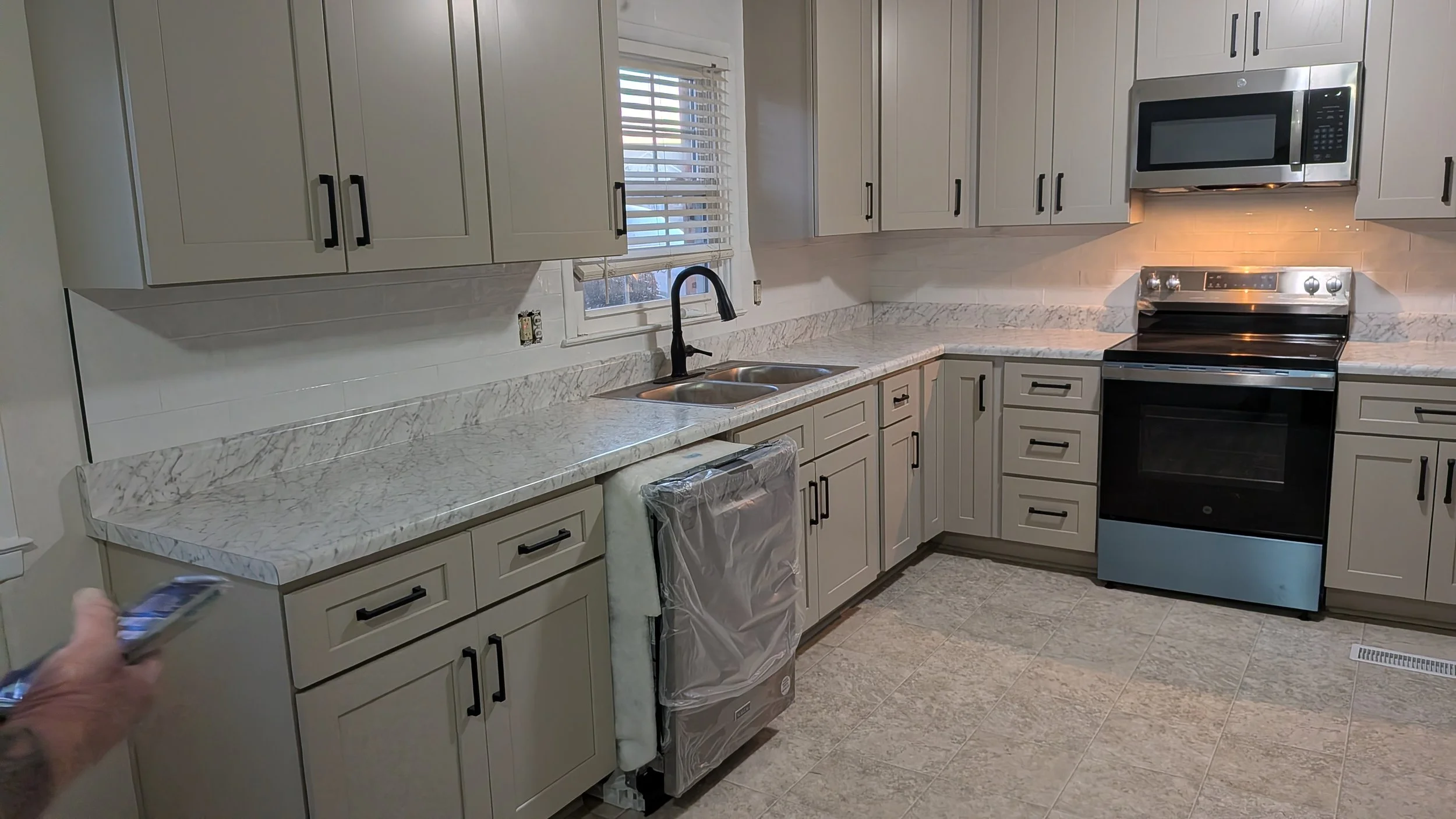 Empty kitchen with white cabinets, granite countertops, black hardware, black appliances, including microwave, oven, dishwasher wrapped in plastic, and a double sink with black faucet, window with blinds above the sink, and tiled floor.