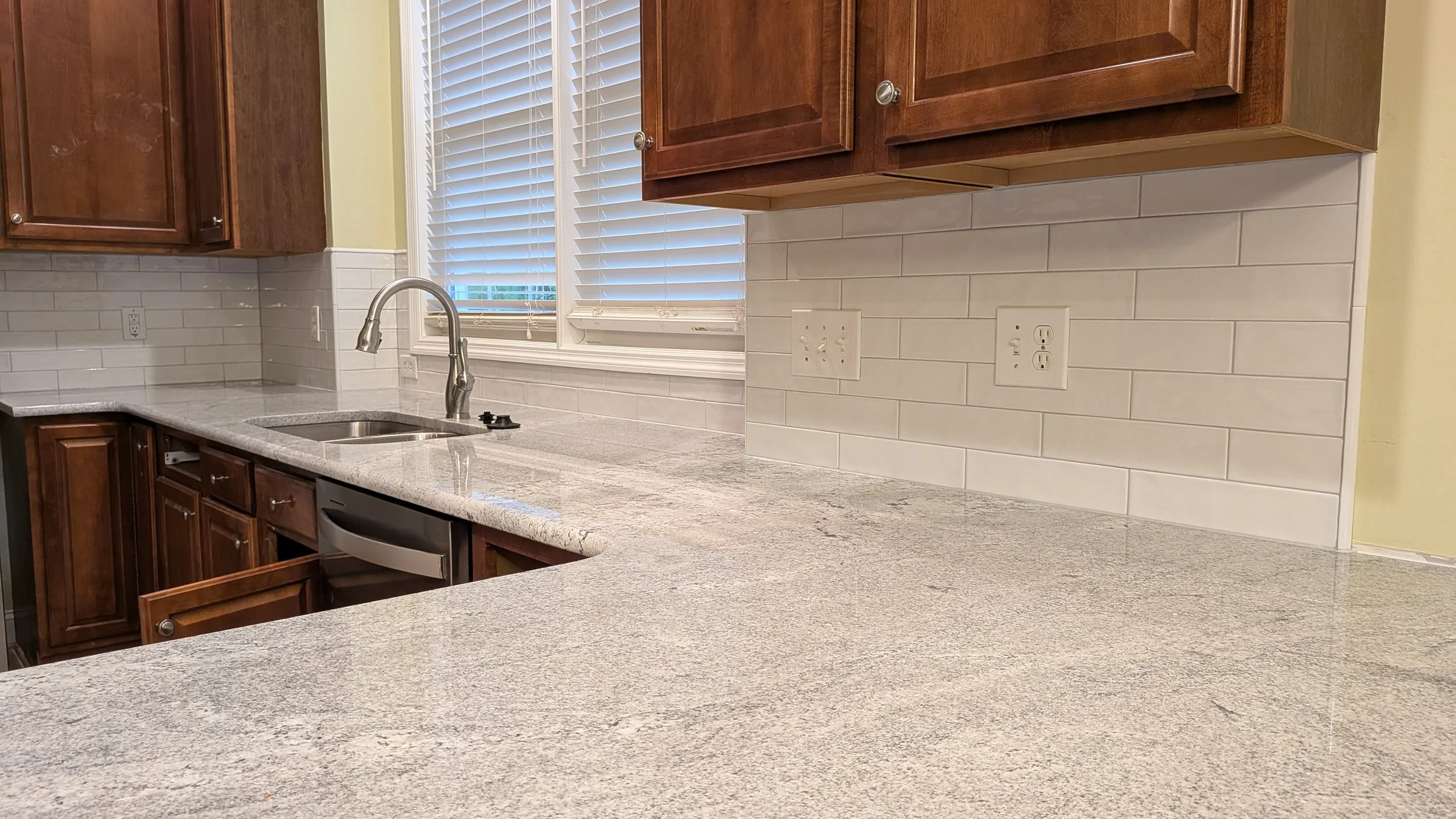 View of a kitchen countertop with granite surface, white subway tile backsplash, a stainless steel sink, and wooden cabinets. There are electrical outlets on the backsplash and a window with blinds above the sink.