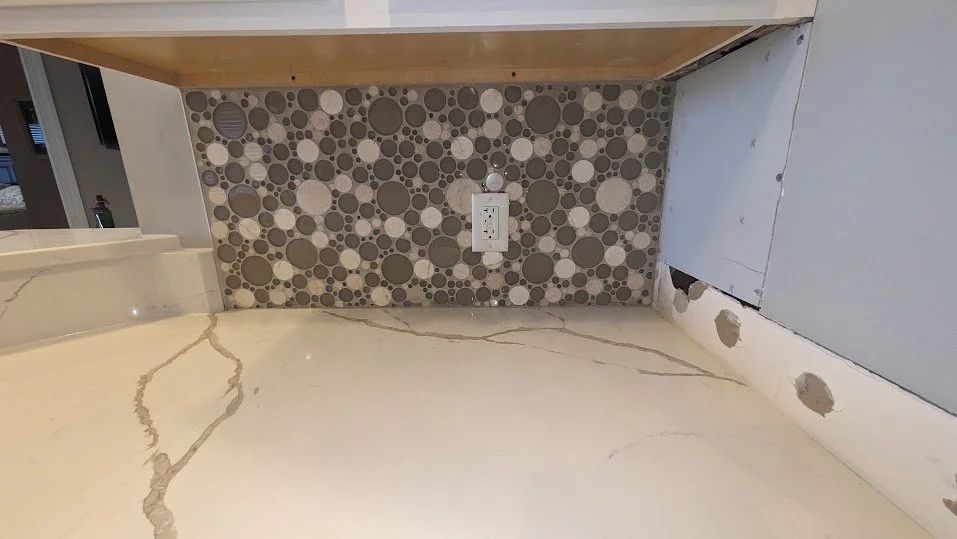 Kitchen backsplash with a circular mosaic tile pattern in shades of gray and beige, an electrical outlet, and part of a marble countertop.