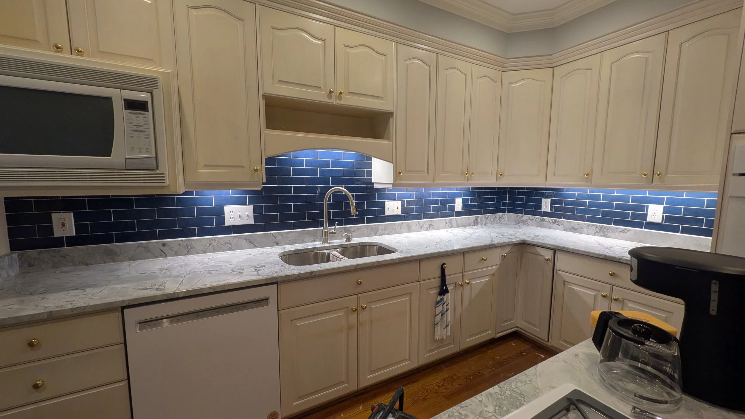 Kitchen with cream-colored cabinets, a marble countertop, dark blue tile backsplash, and a window over the sink. Appliances include a microwave, dishwasher, and coffee maker.