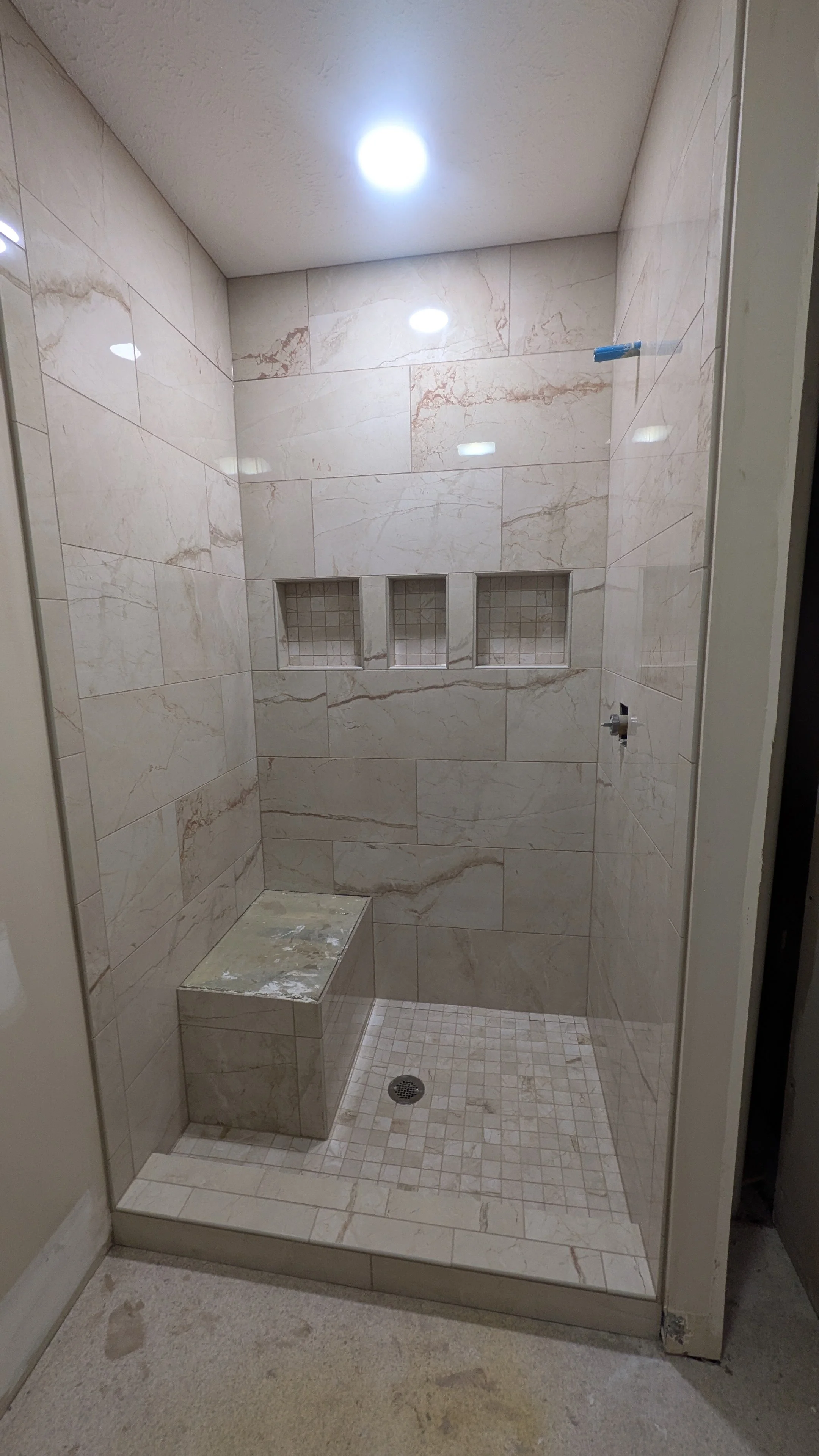 A tiled shower with three recessed wall shelves, marble tiles, a built-in bench, and a floor drain, under a ceiling with two recessed lights.