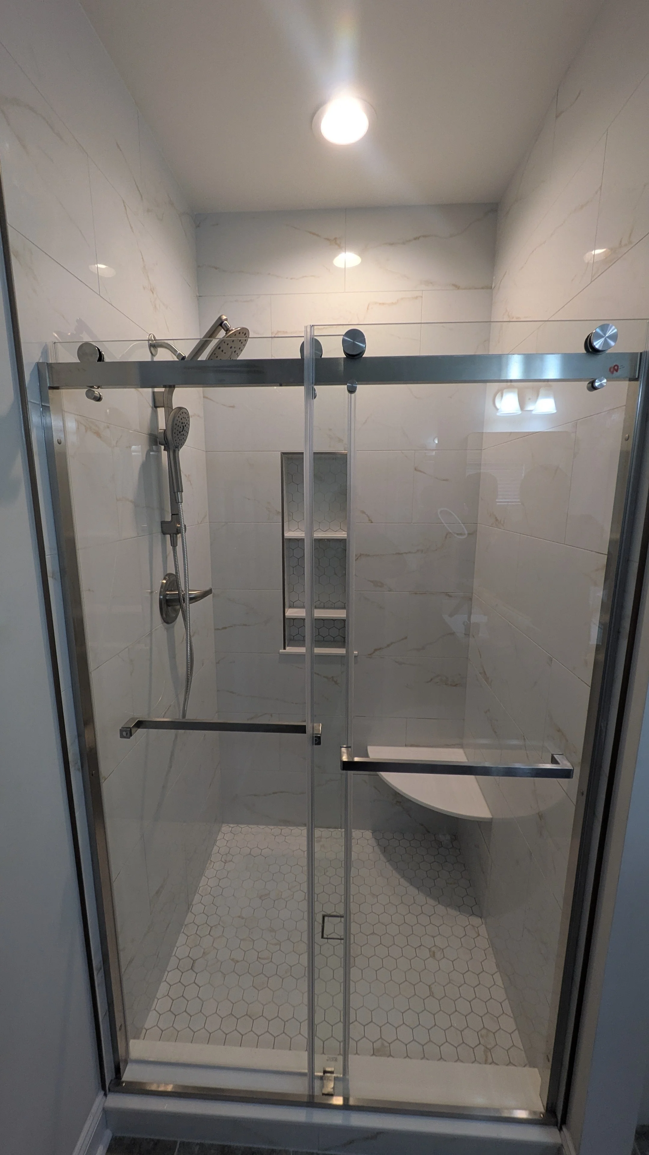 A modern walk-in shower with glass sliding doors, marble-like wall tiles, a built-in niche with shelves, a bench, and multiple showerheads.