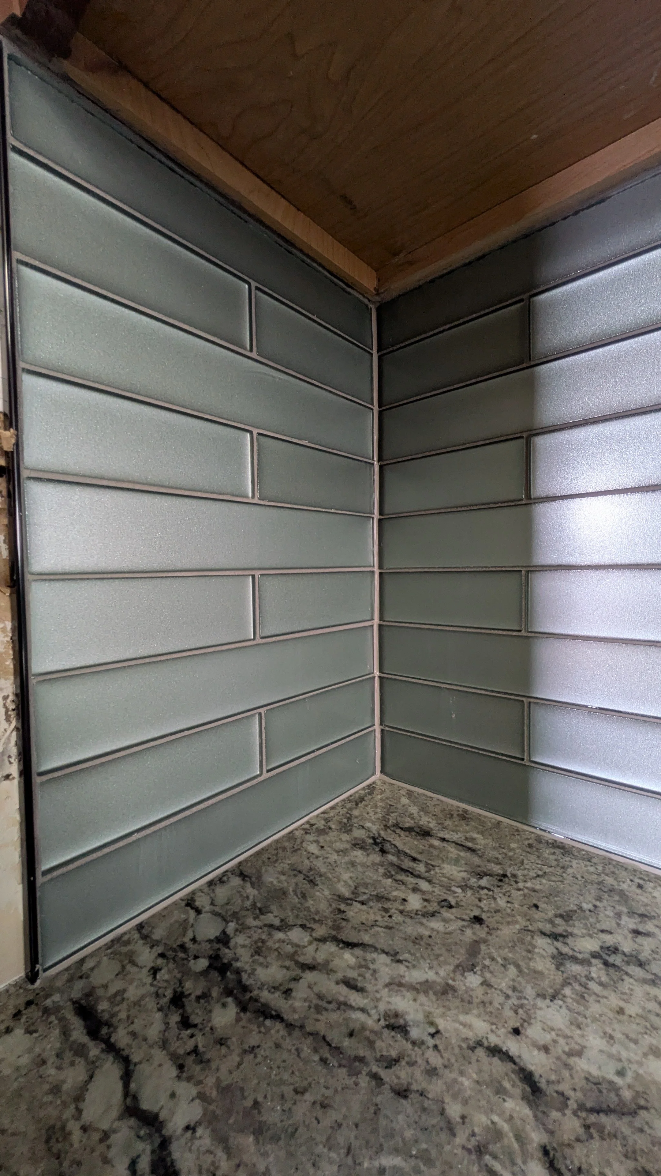 Close-up of a corner of a kitchen countertop with a glass tile backsplash made of rectangular, frosted glass tiles in shades of gray. The countertop is a speckled granite surface, and a wooden cabinet is above.