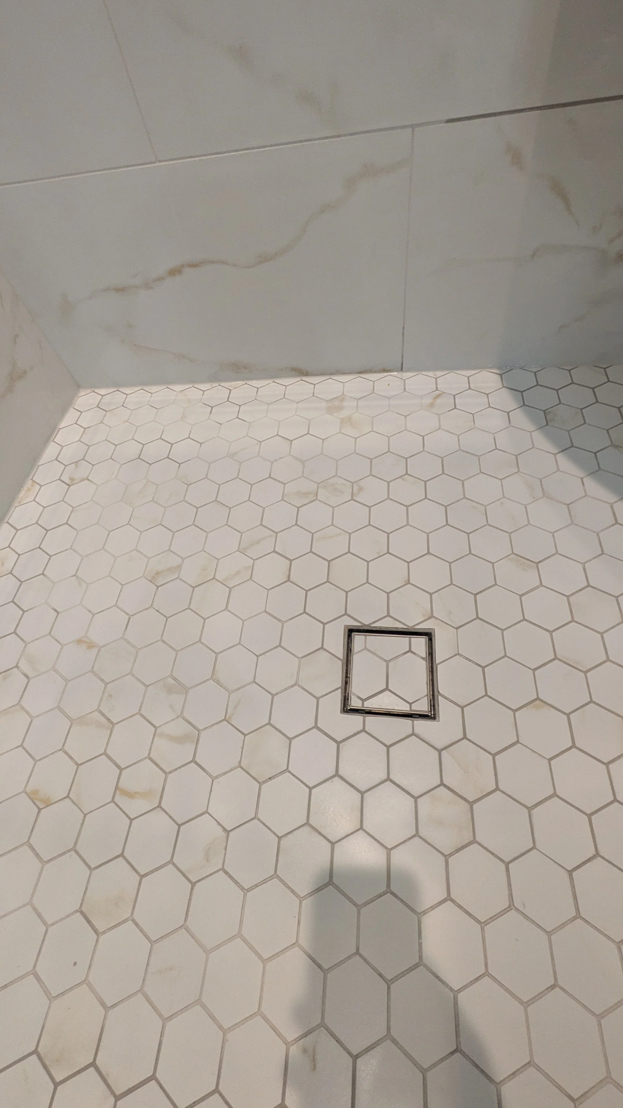 Close-up of a tiled shower floor with white hexagonal tiles, a square drain, and marble-patterned wall tiles.