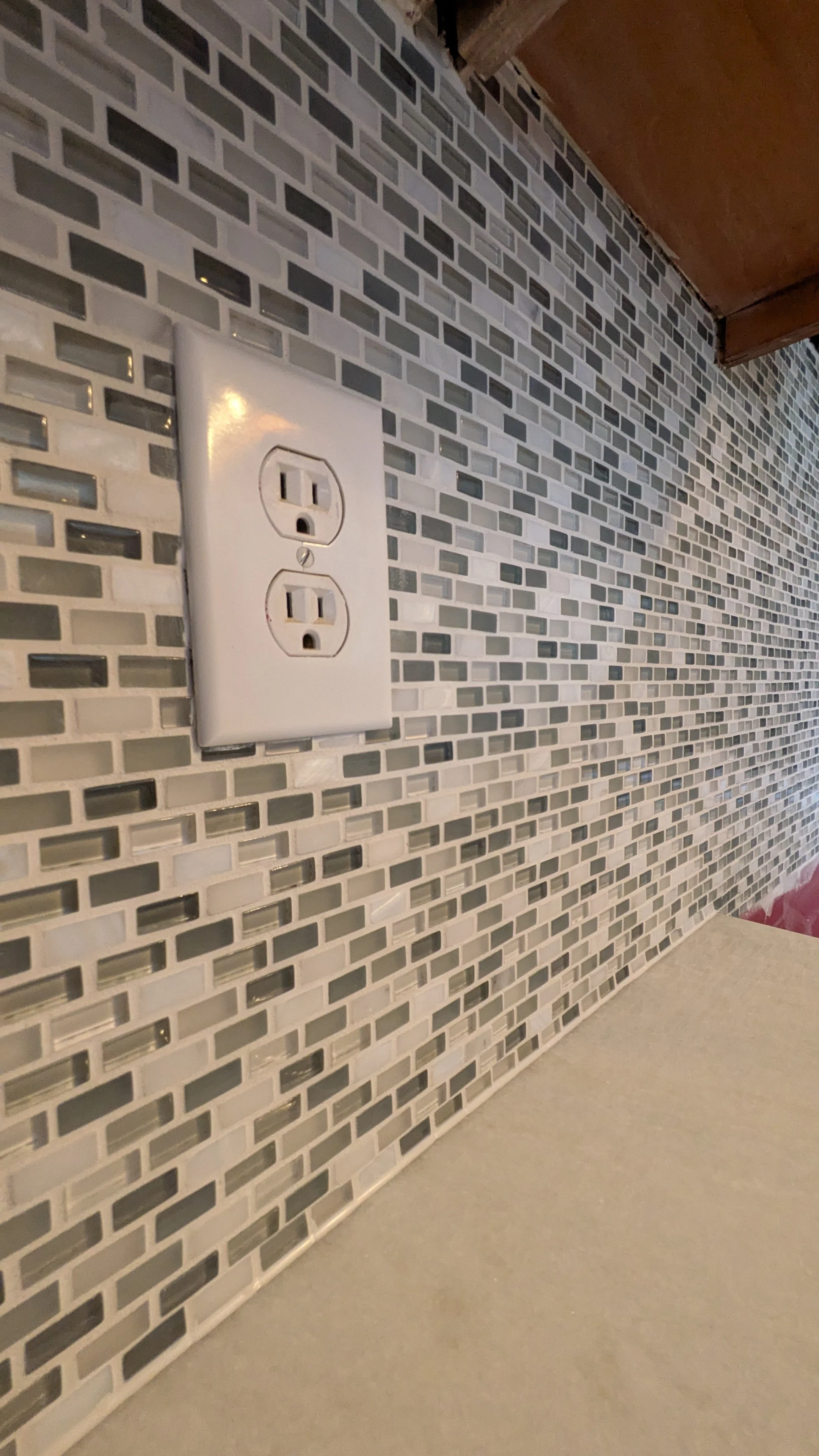 A close-up of a white double electrical outlet on a gray and white mosaic tile backsplash, with a beige countertop in the foreground and a wooden cabinet above.