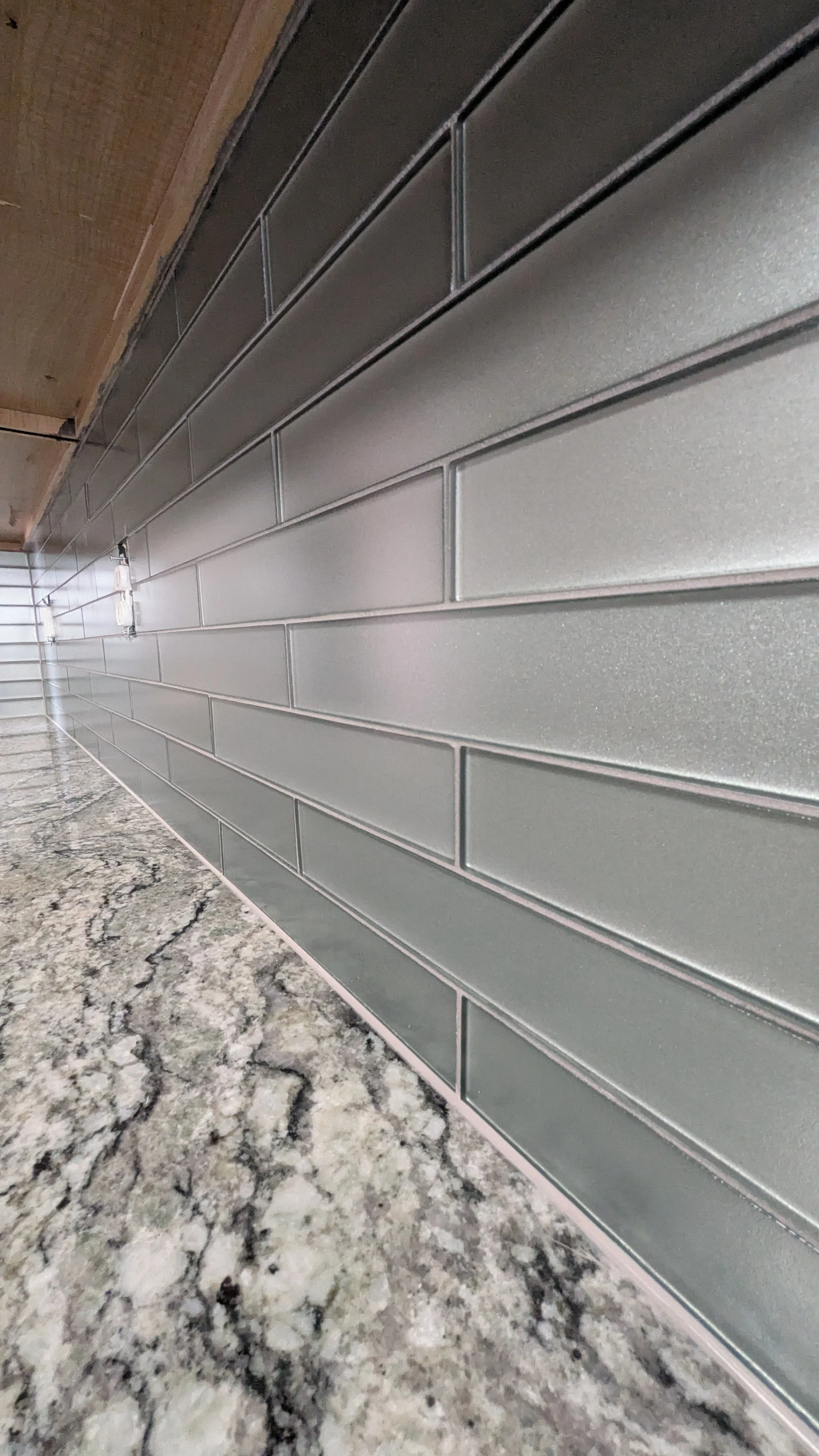 Close-up perspective of a kitchen countertop with gray granite, wooden cabinet, and gray subway tile backsplash.