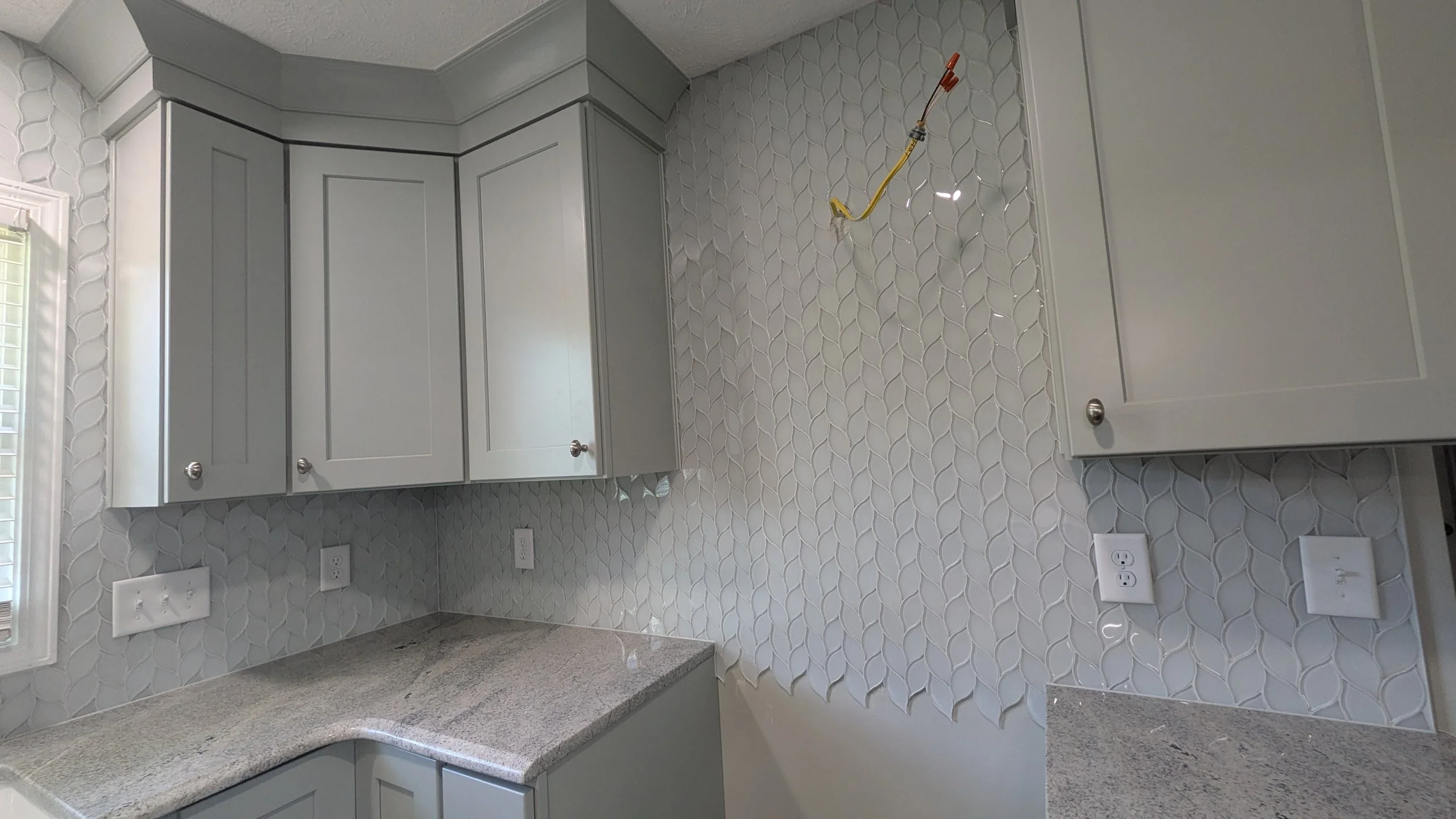 Kitchen with light gray cabinets, a granite countertop, and a decorative tile backsplash. There are electrical outlets and an open cabinet with a wire hanging from the ceiling.