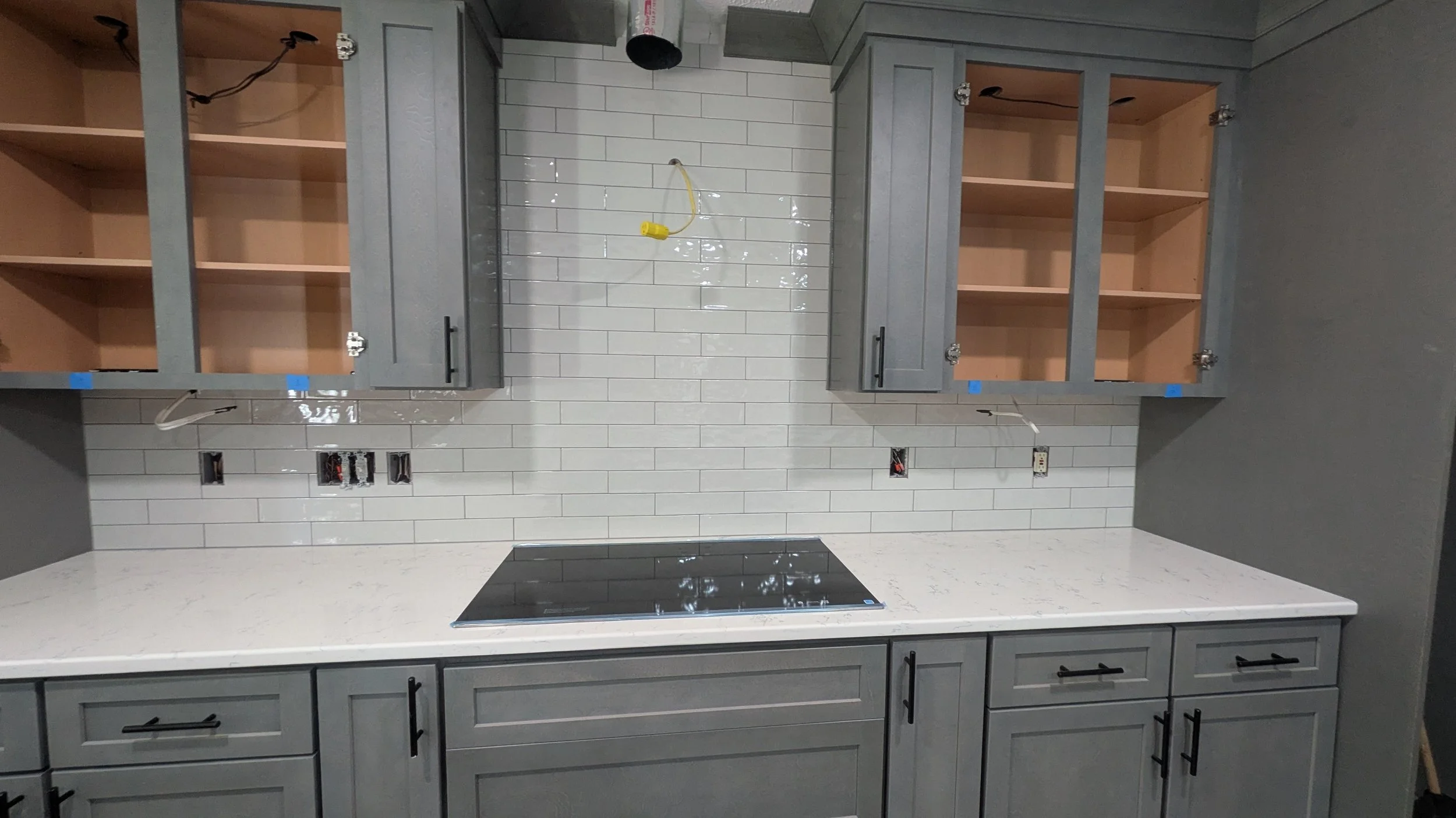 Kitchen with gray cabinets, white subway tile backsplash, and a black cooktop on a white countertop, some electrical outlets and missing cabinet doors.