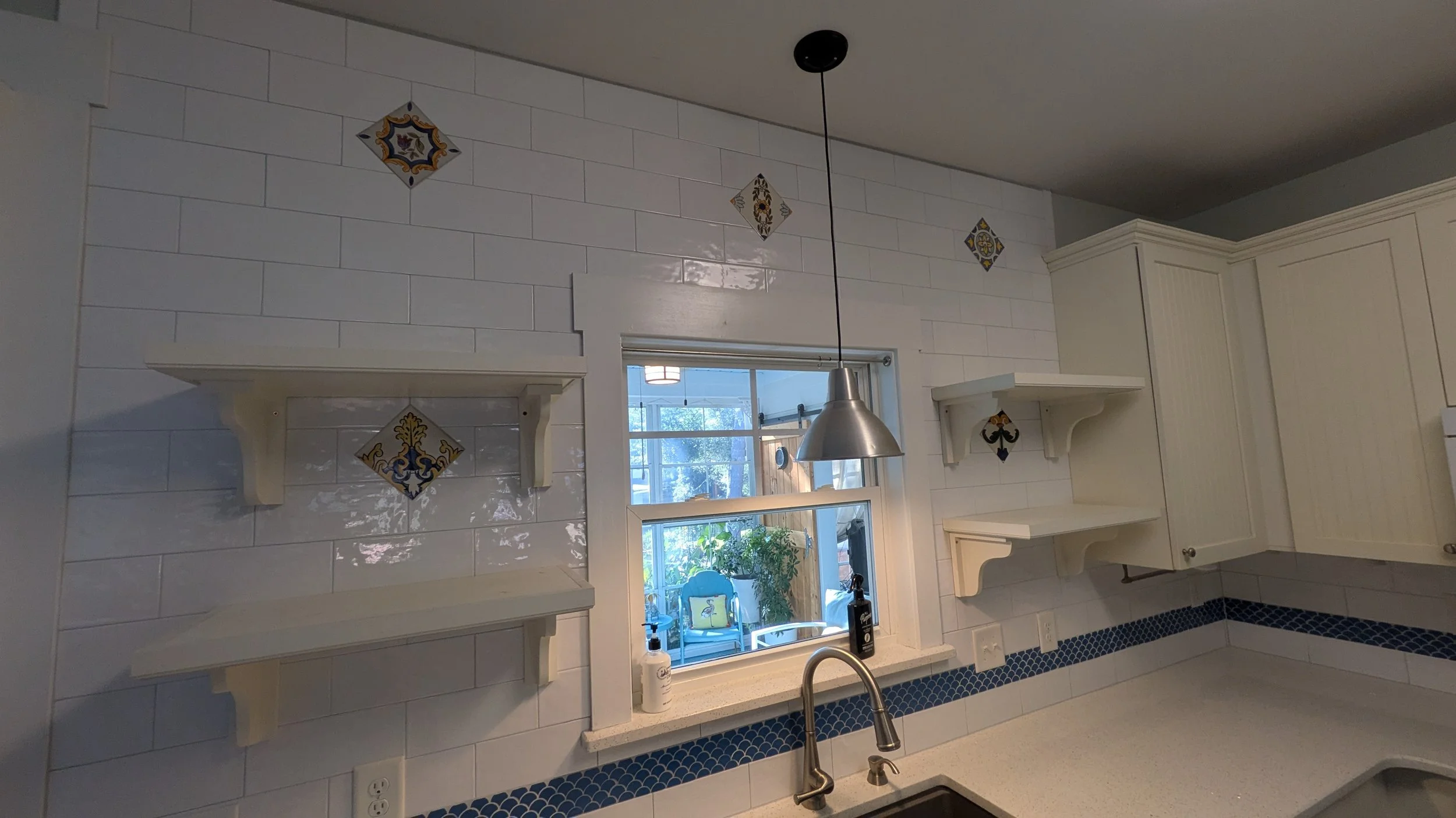 Kitchen with white cabinets, decorative tiles, open shelves, a window with a view of a porch and outdoor furniture, and a hanging light fixture.