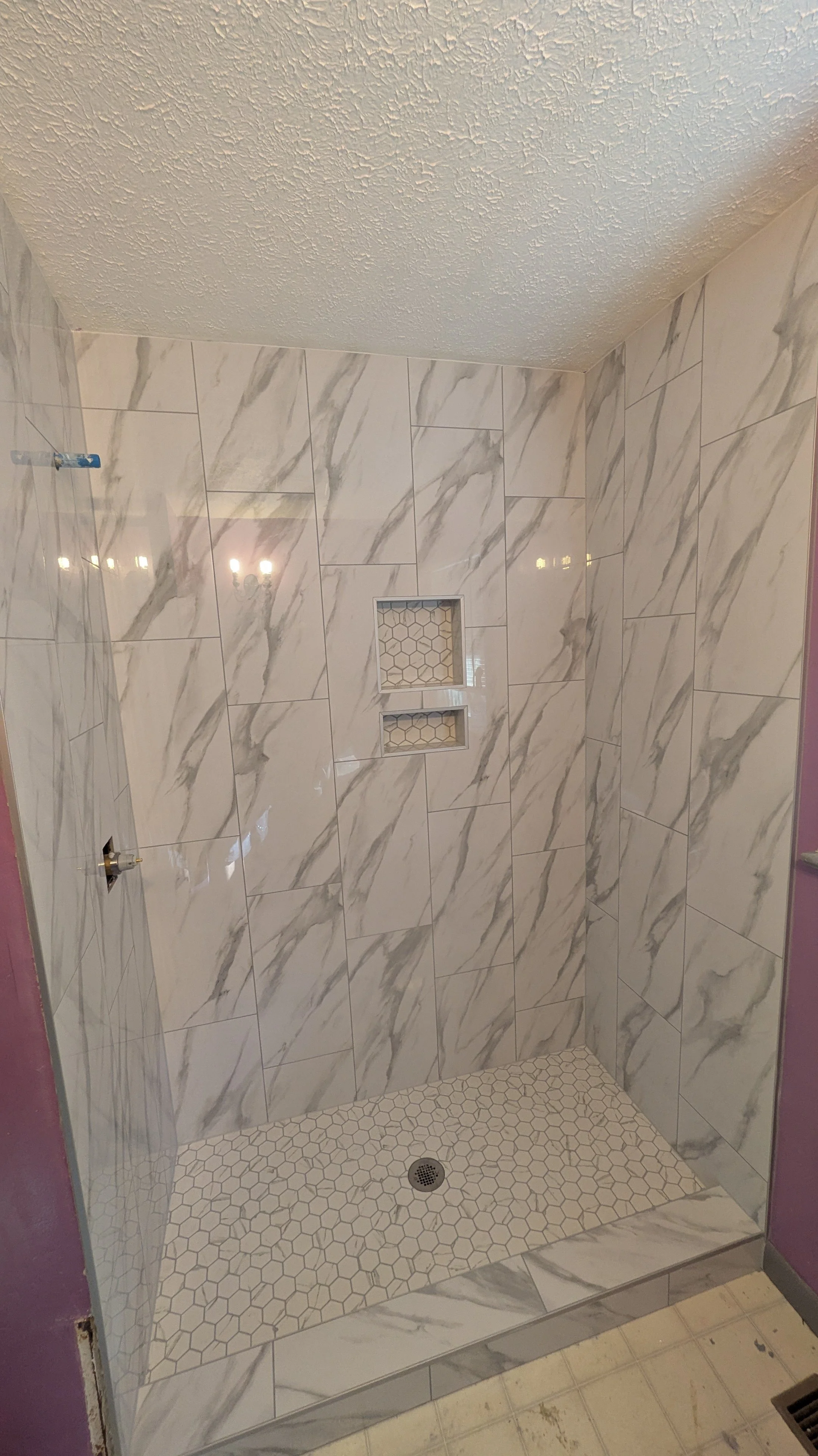 A newly renovated walk-in shower with marble wall tiles, two recessed shelves with honeycomb tile patterns, a drain at the center of a small hexagon tile floor, and purple painted partial walls on each side.