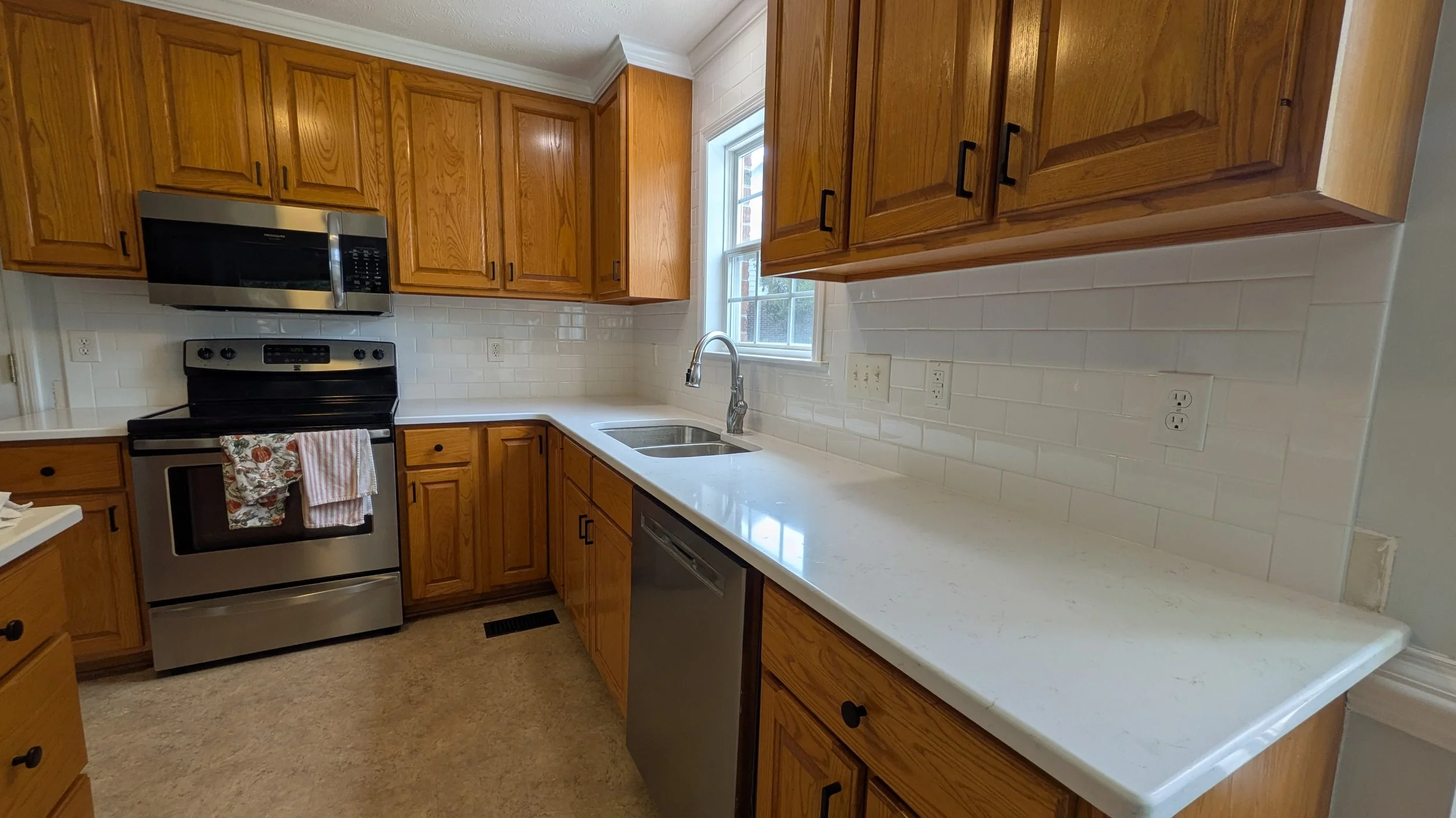 Kitchen with wooden cabinets, stainless steel microwave and oven, white countertop, double sink, and beige carpeted floor.