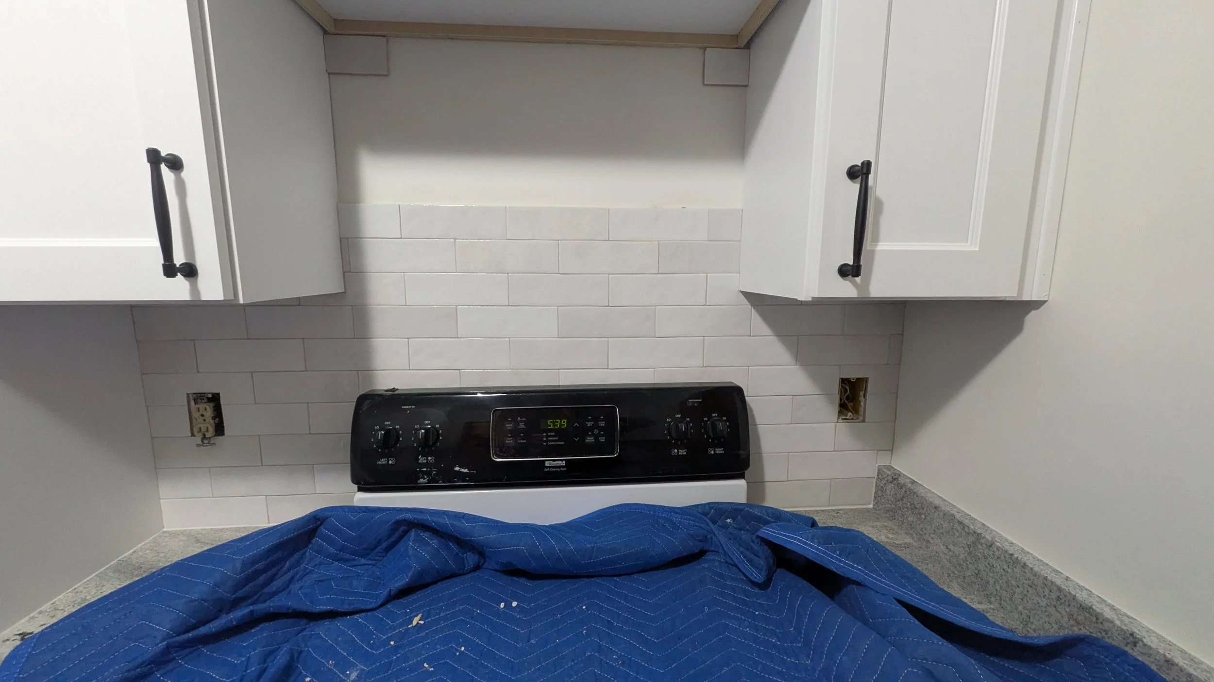 Kitchen with white cabinets, a black electric stove, and a gray countertop. The wall behind the stove has white subway tiles, with two electrical outlets and one missing cover plate.