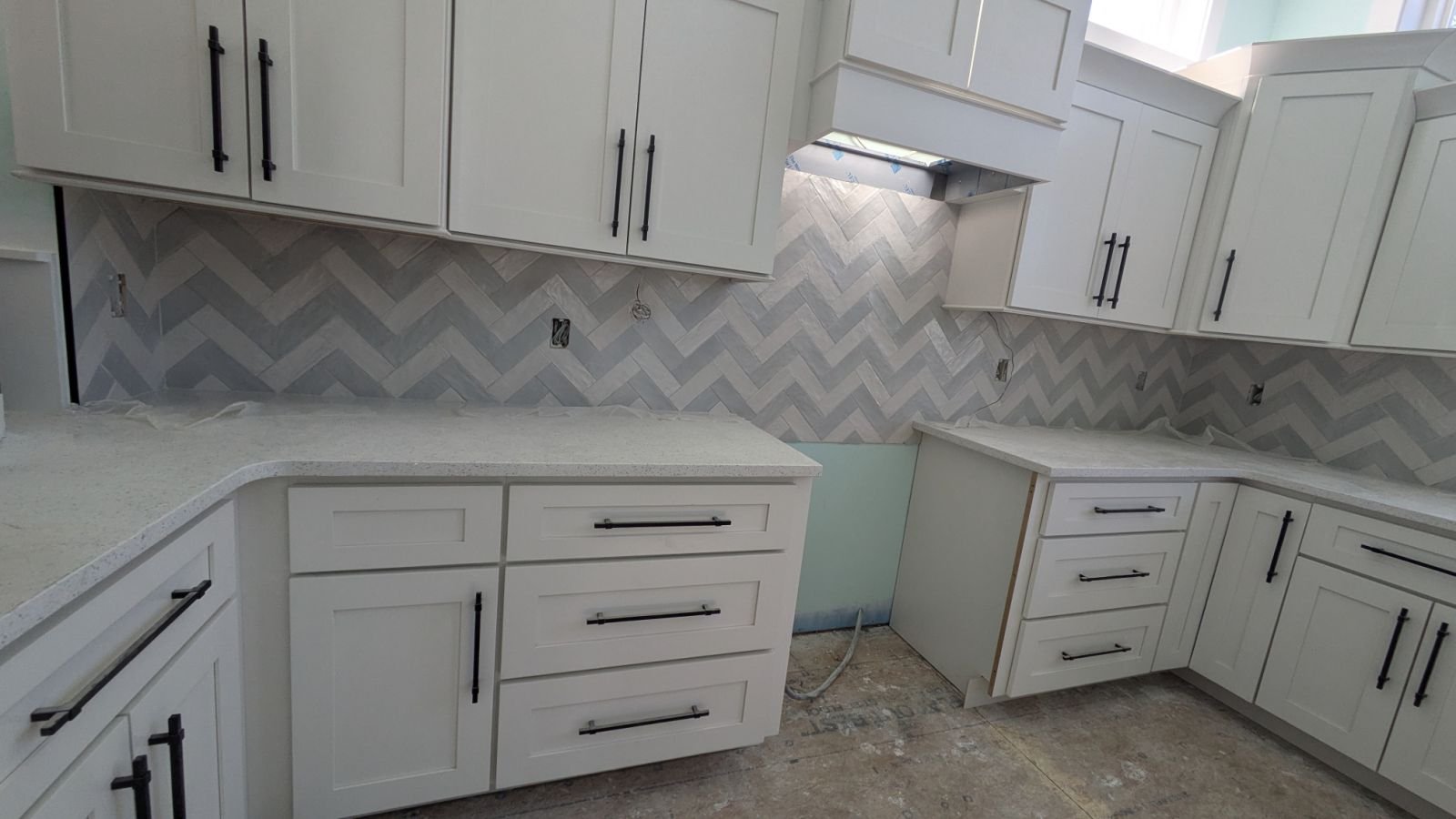 Kitchen with white cabinets and black handles, gray and white herringbone tile backsplash, and unfinished flooring.