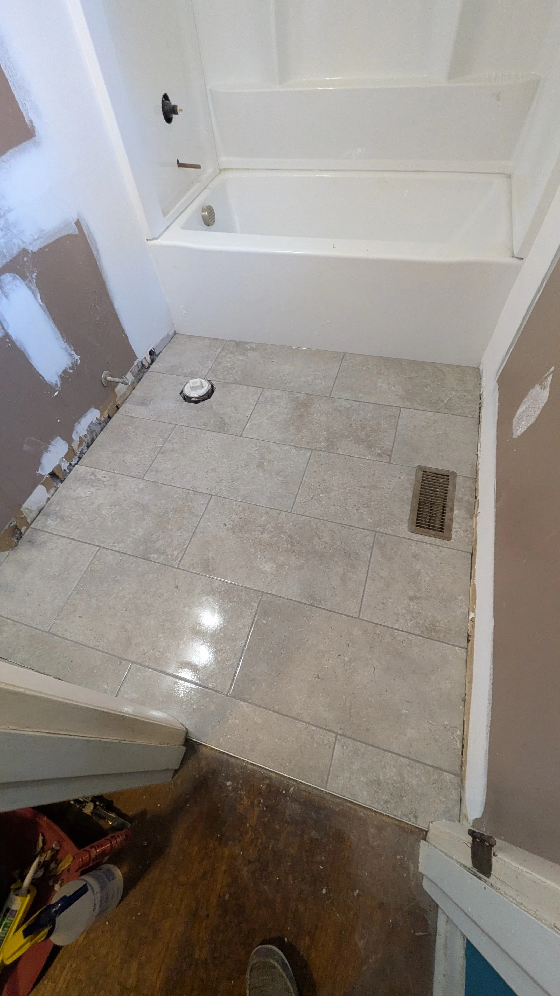 Bathroom floor with beige tiles, a bathroom shower with white walls, plumbing fixtures, a drain, and construction tools and materials nearby.