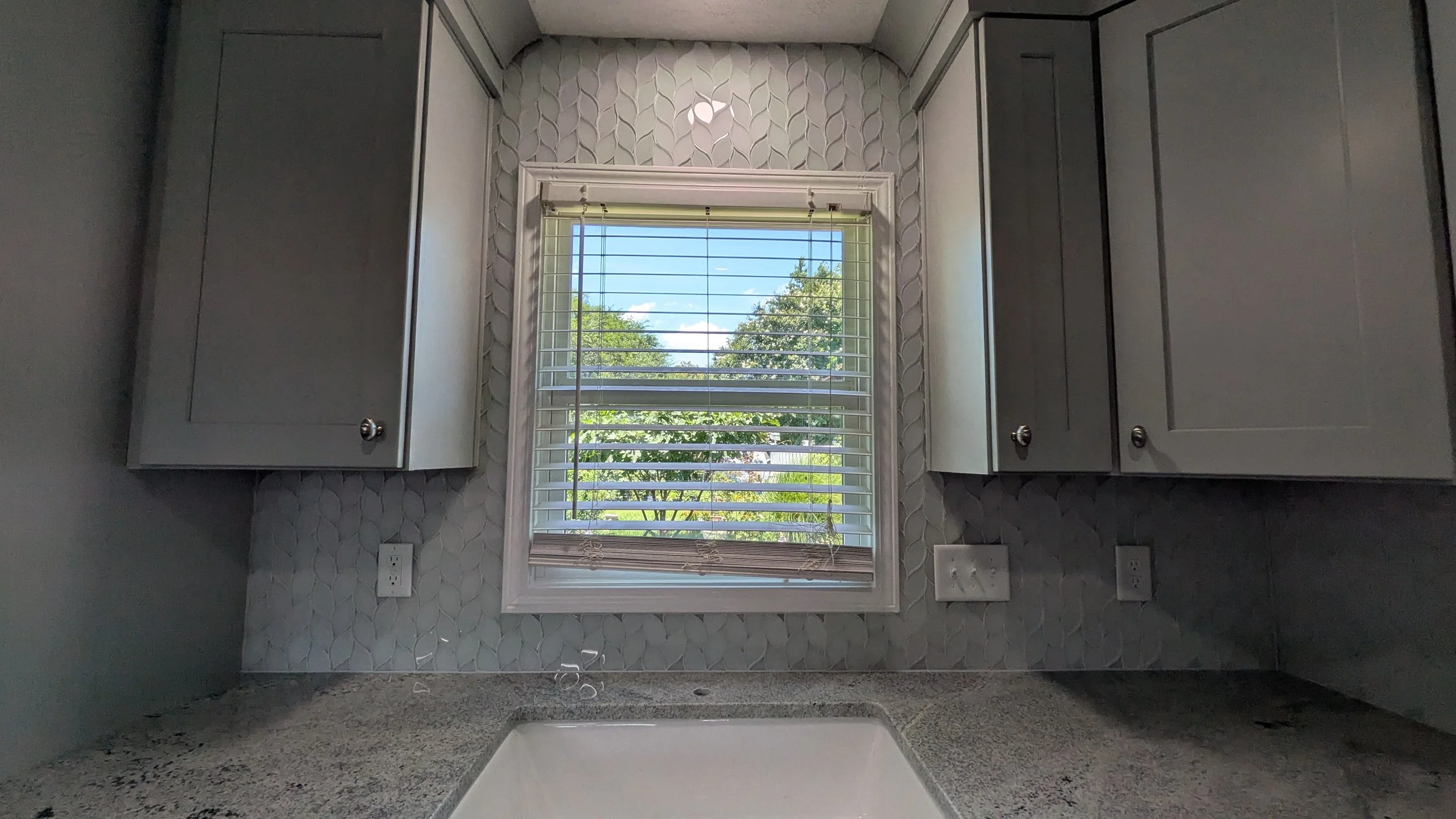 Kitchen window with blinds above a sink, with gray cabinets on either side and a textured backsplash and countertop.