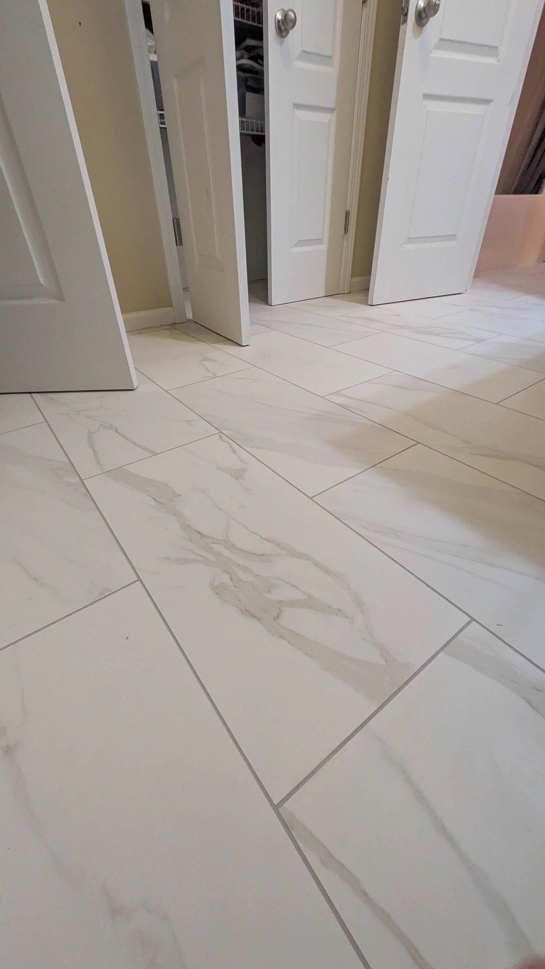 View of a room with a white tiled floor featuring beige and light brown veining, with open closet doors revealing clothes inside.