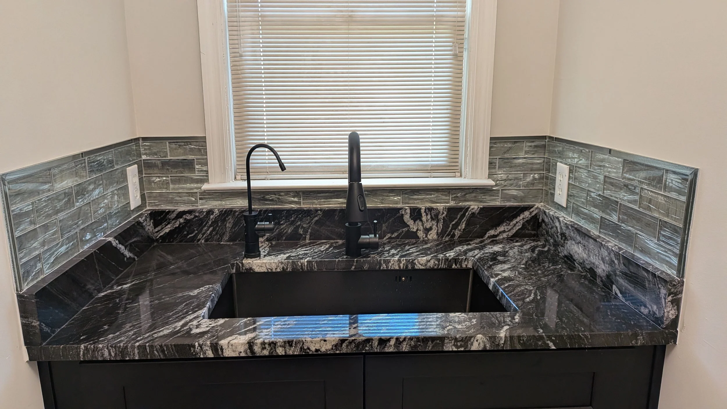 Black granite kitchen sink with a high arc black faucet, a matte black soap dispenser, and a window with closed blinds above the sink, surrounded by glass tile backsplash and electrical outlets.