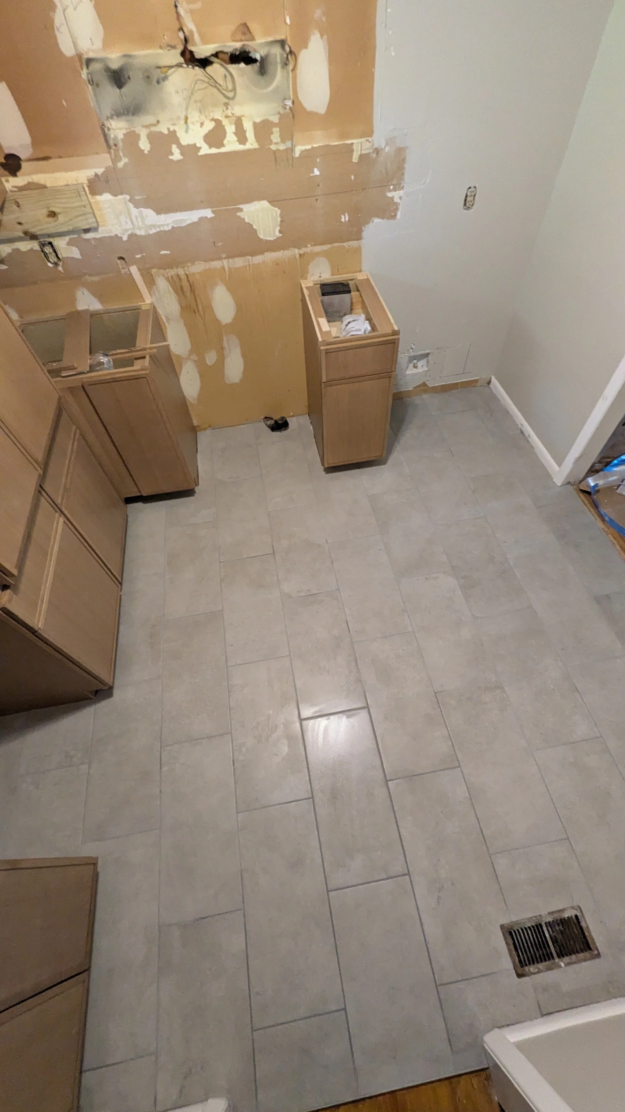 Kitchen under renovation with partially installed beige tile flooring, unfinished wall behind cabinets, and plumbing fixtures exposed.