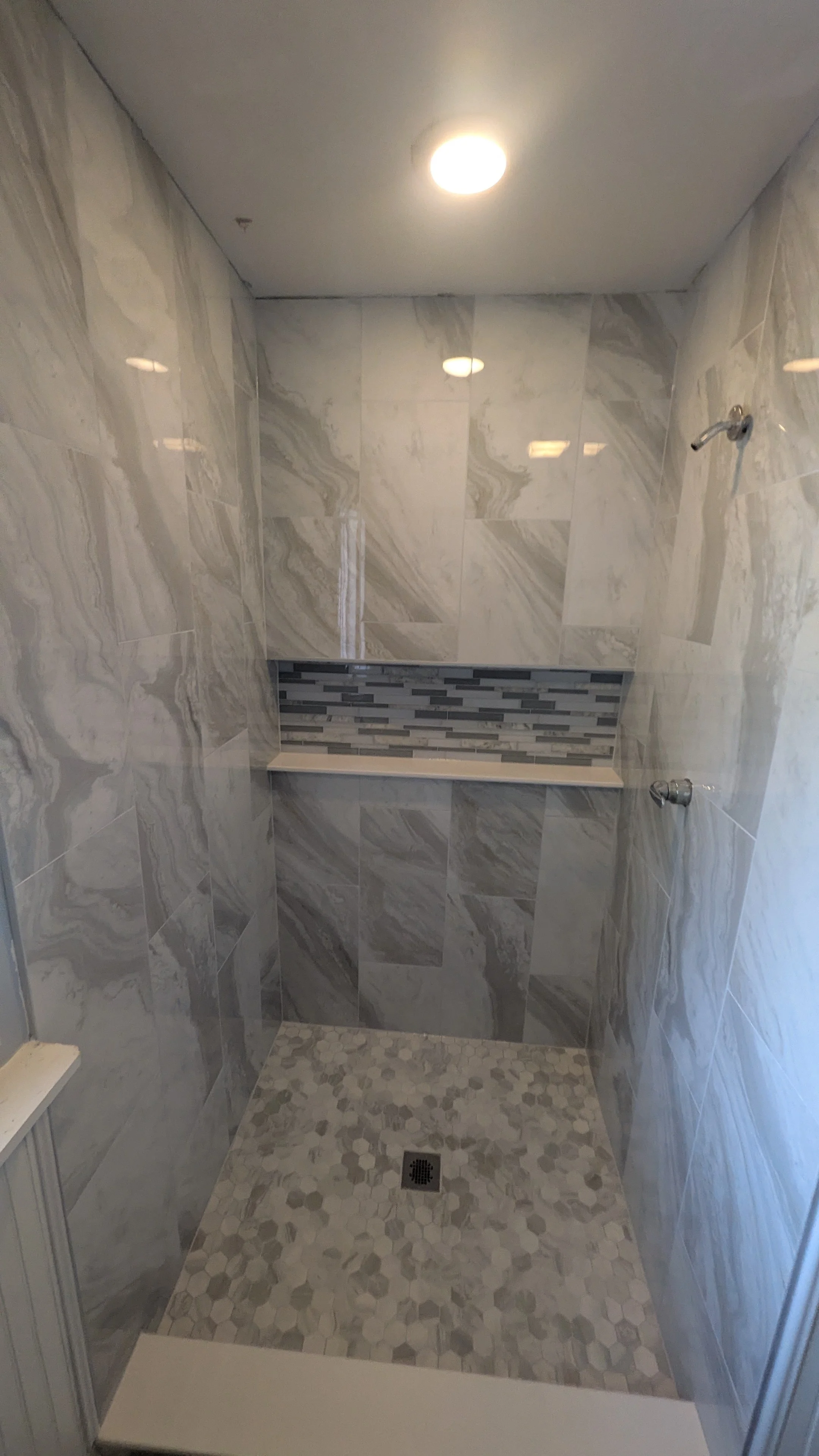 A walk-in shower with gray marble-patterned tiles, a strip of gray and black mosaic tiles on the back wall, and a built-in shelf. The shower also has two hooks on the side wall and a drain on the floor. There is a ceiling light illuminating the space