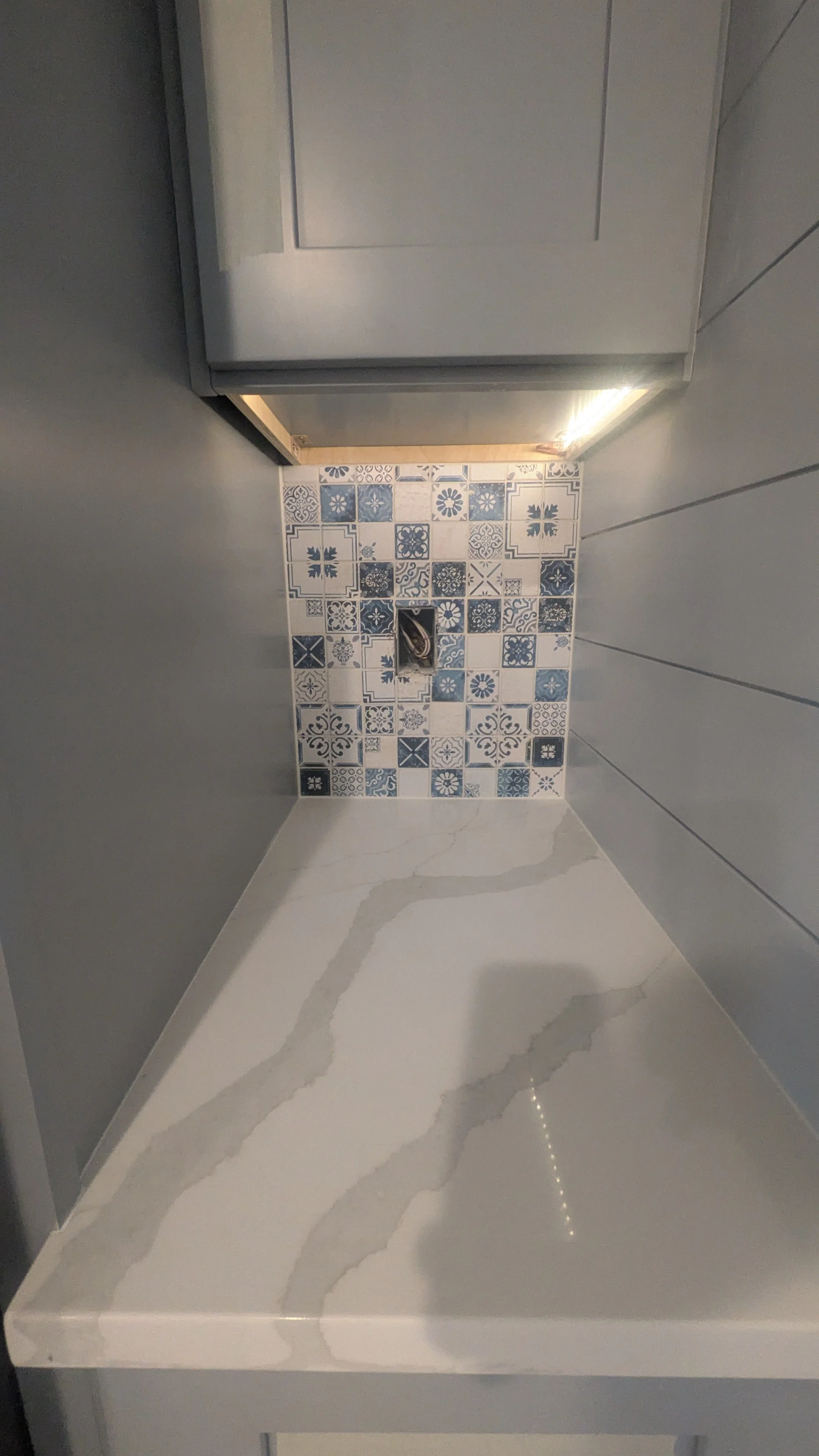 Close-up of a kitchen countertop with white marble design and gray veins, a back wall with blue and white patterned tiles, and a gray cabinet above.