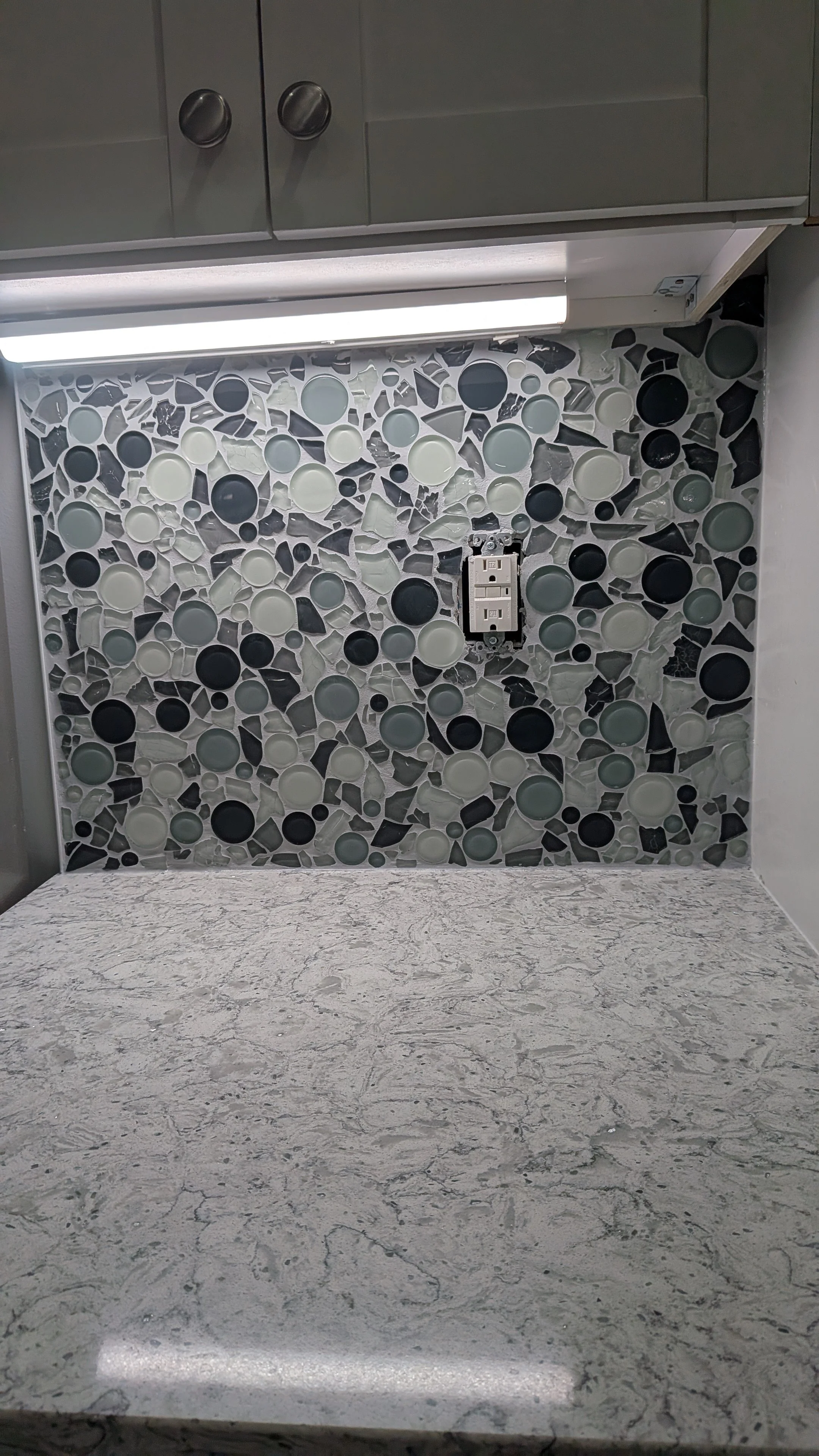 Kitchen backsplash with black, white, and gray mosaic tiles and a countertop with a gray and white marbled pattern. An electrical outlet is mounted on the backsplash. Kitchen cabinets above with knobs and under-cabinet lighting.