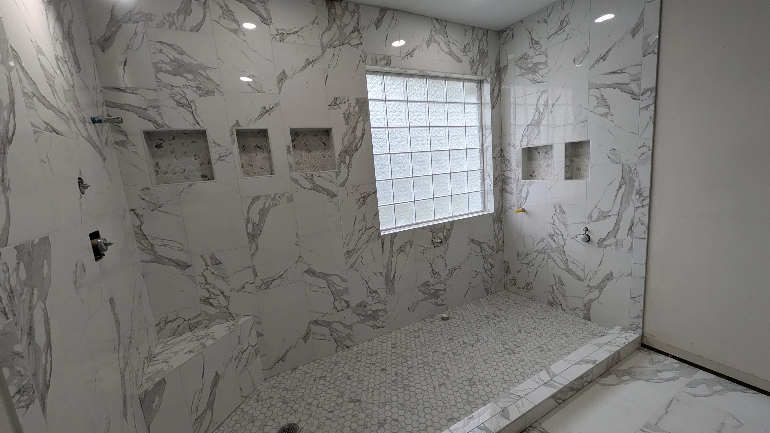 Empty shower area with marble-like tile walls and small hexagonal floor tiles, with built-in shelf niches and a window.
