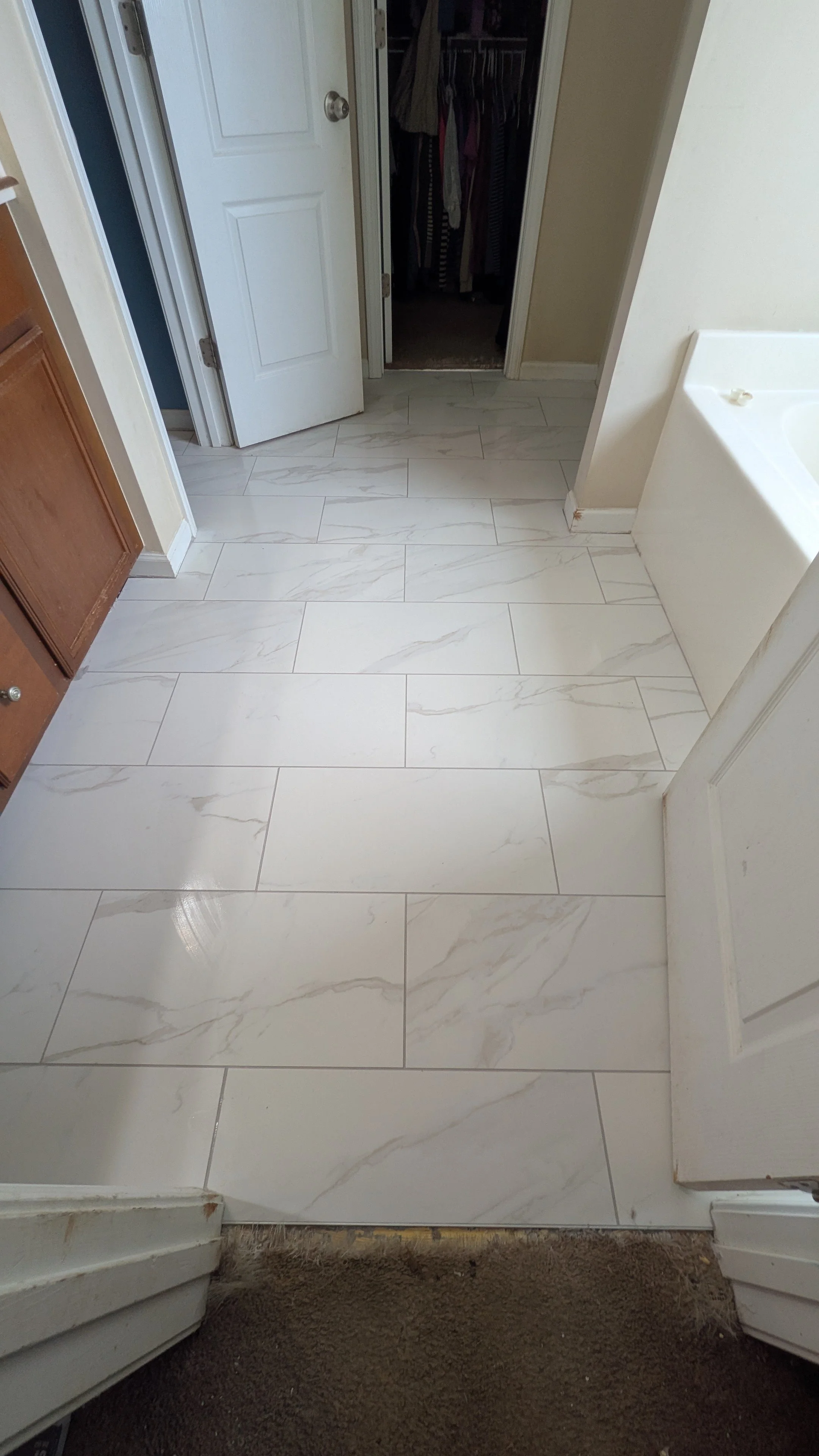 Bathroom with white marble floor tiles, wooden cabinets, a white bathtub, an open closet filled with clothes, and a toilet area in the background.