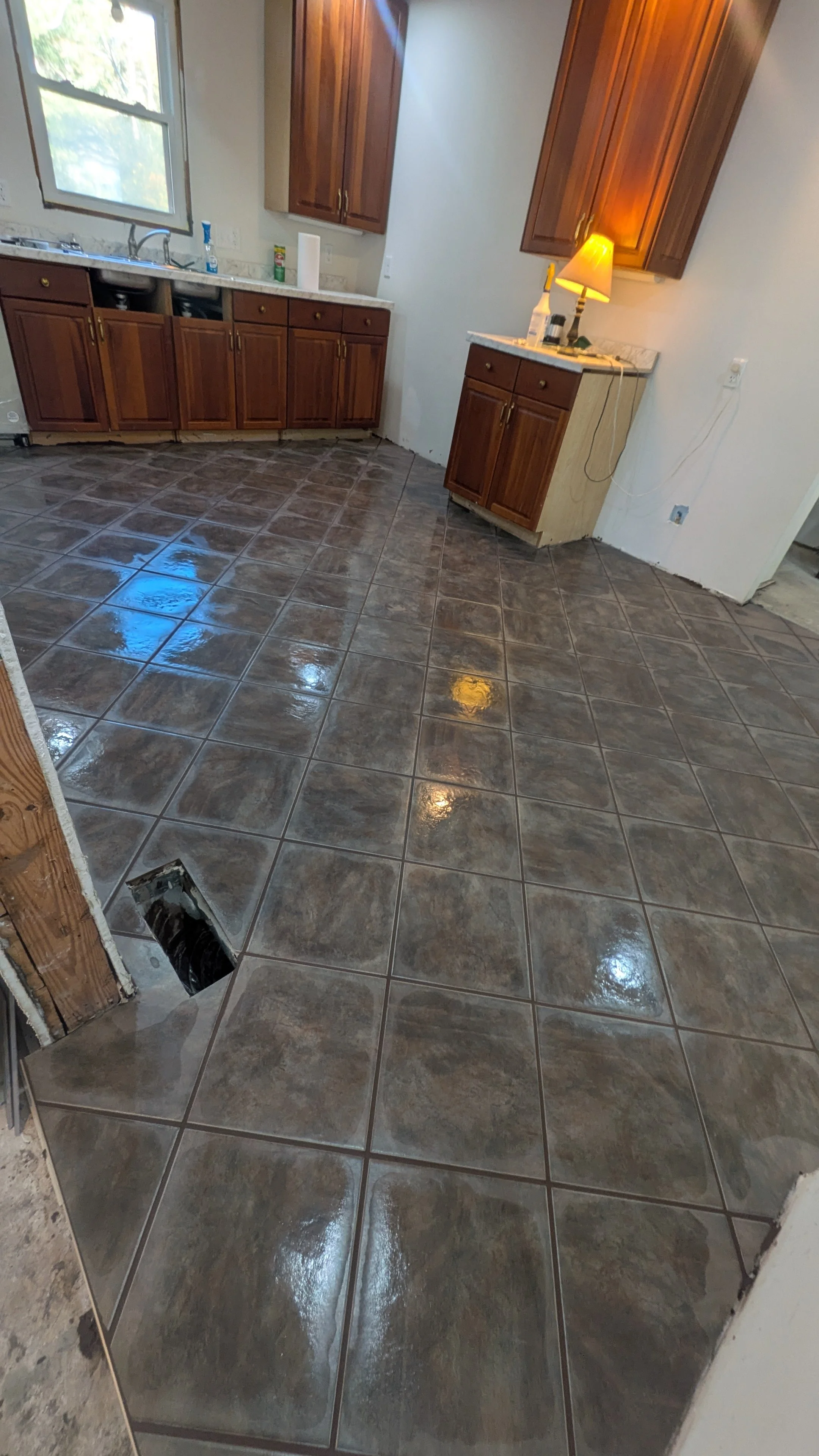 Kitchen with brown wooden cabinets, white countertops, a window above the sink, a yellow lamp on a small cabinet, and new glossy brown floor tiles.
