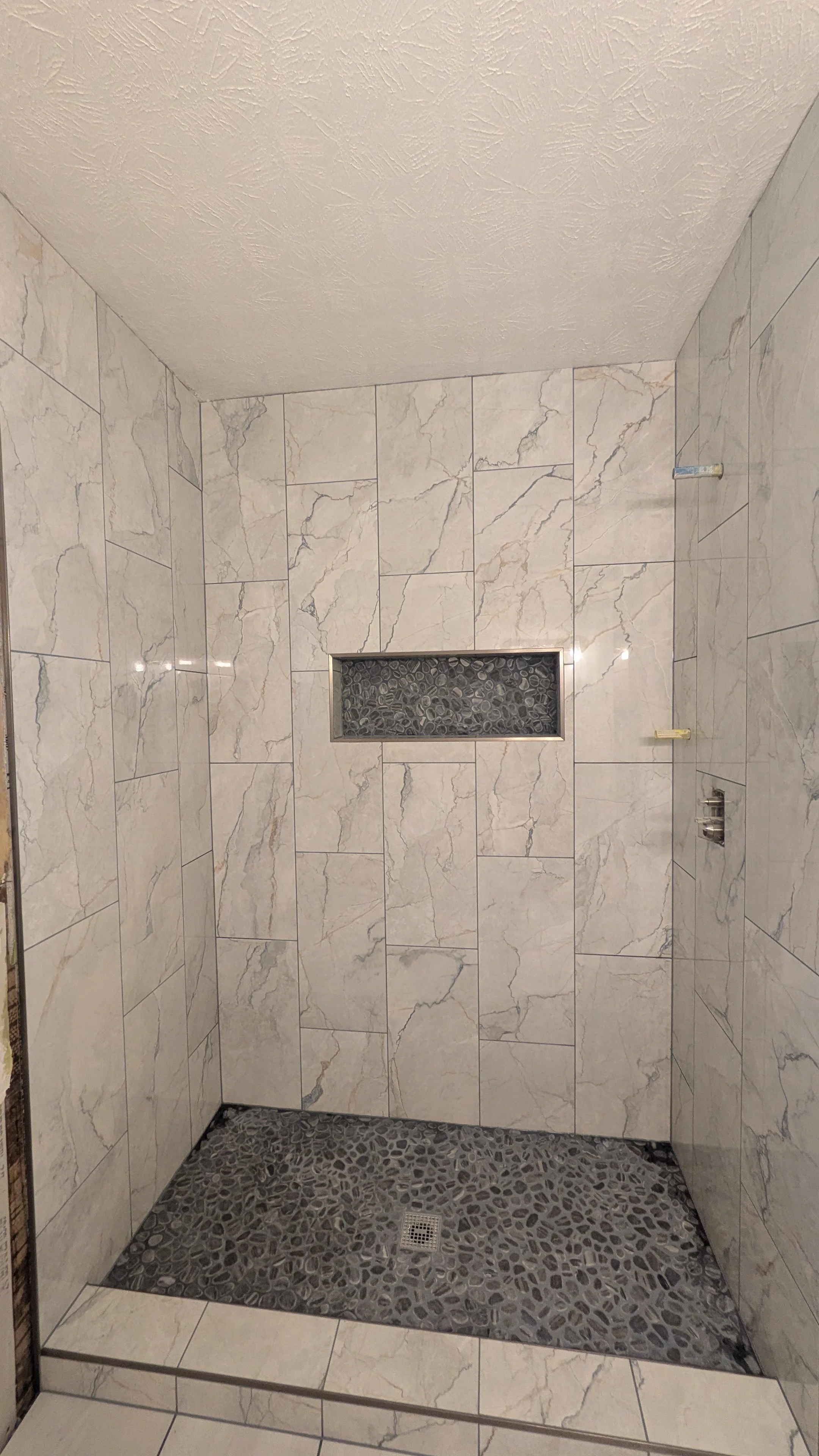 Modern shower with marble tiles on walls, pebble stone on floor, a small niche with pebble stones, and a drain.