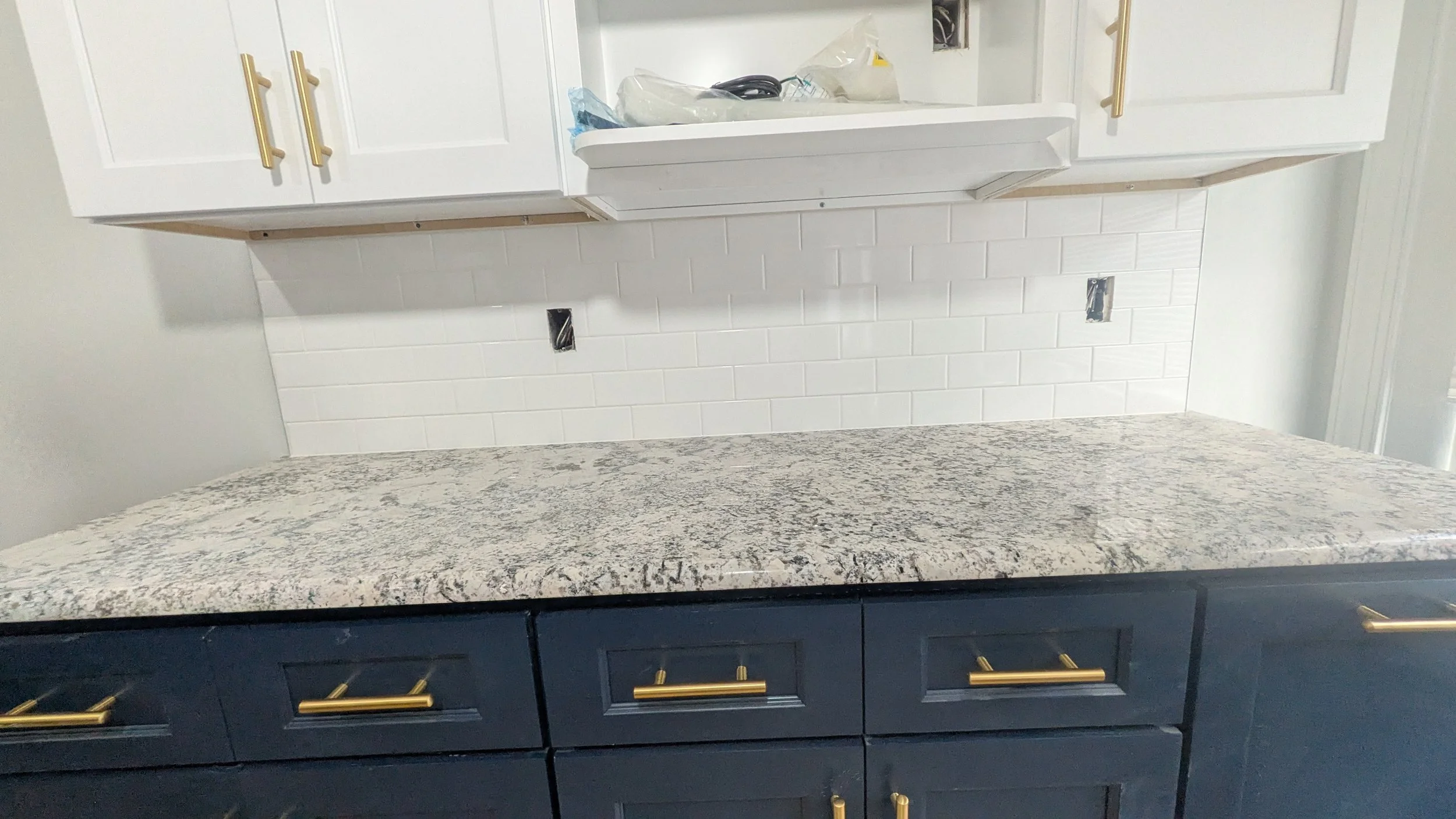 Kitchen countertop under construction, granite surface installed over dark blue cabinets with gold handles, white subway tile backsplash with electrical outlets, and white upper cabinets with gold handles.