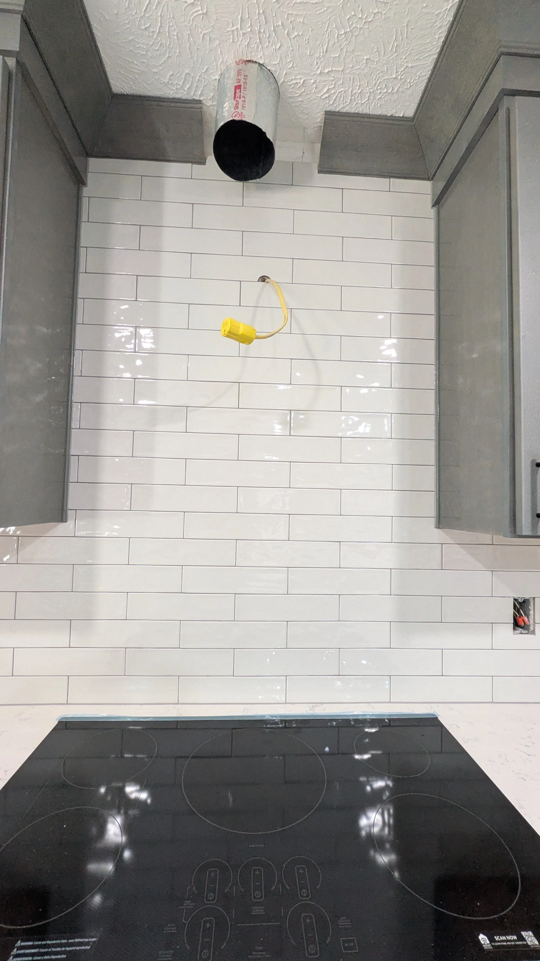 Empty kitchen with a white subway tile backsplash, gray cabinets, a black electric stovetop, and a ceiling light fixture. An electrical outlet is visible near the bottom right.