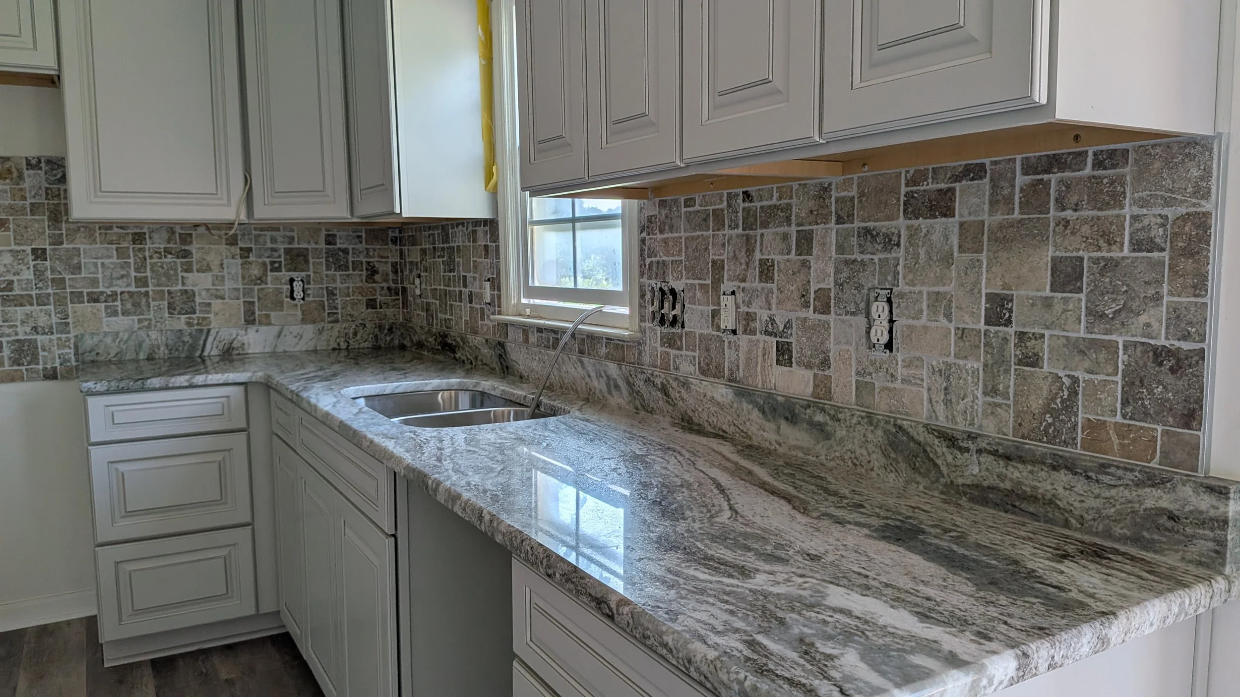 Kitchen counter with granite surface, white cabinets, brick-style backsplash, and a window above the sink.
