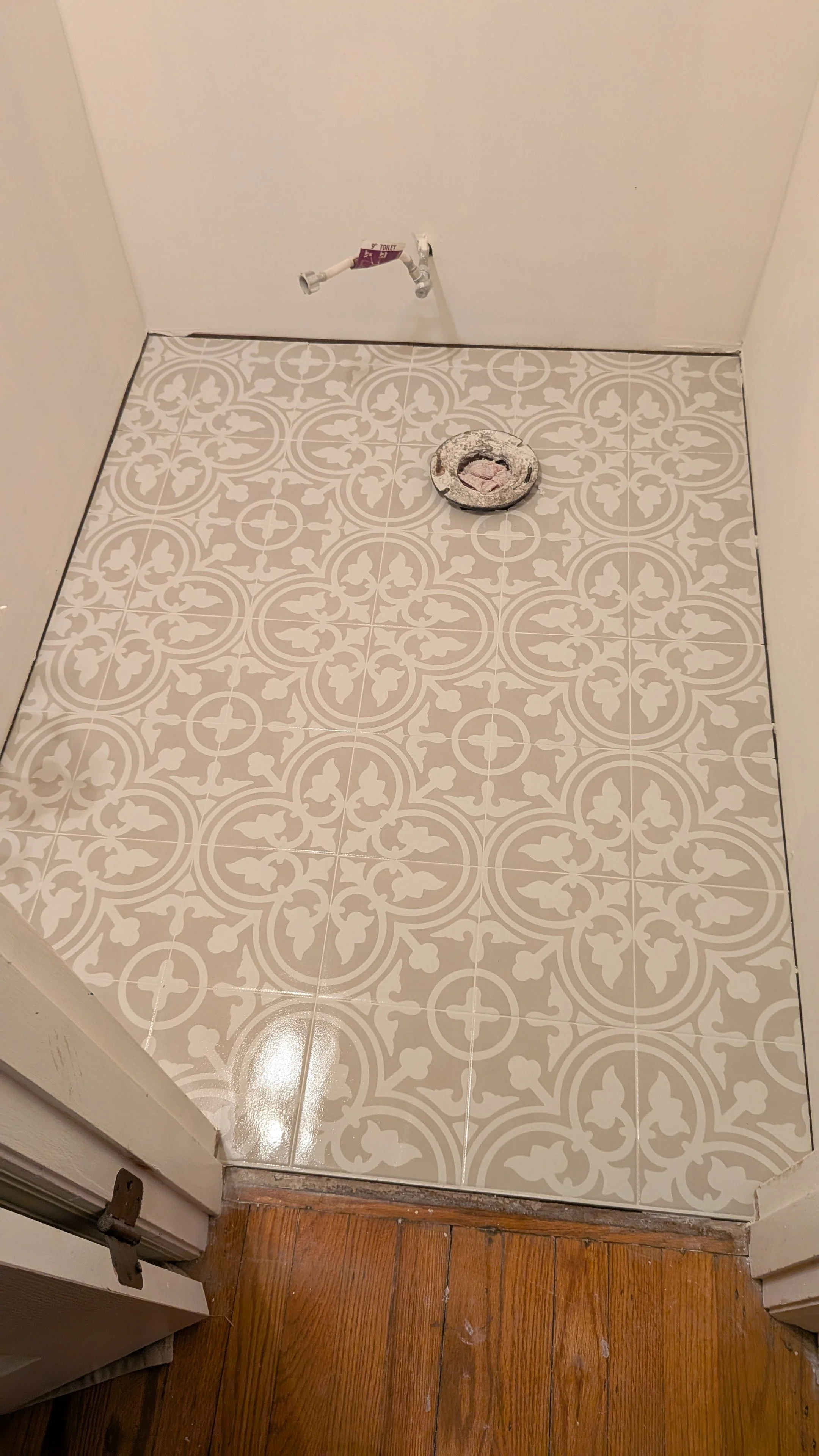 Empty bathroom area with decorated floor tiles, a pipe protruding from the wall, and a round object on the floor.