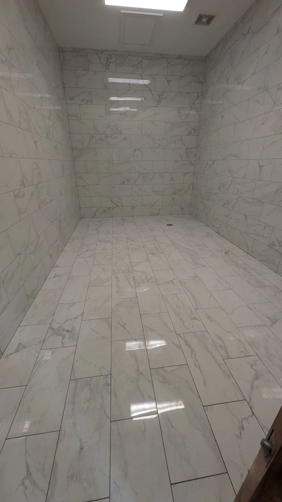 Empty tiled shower with marble-patterned walls and floor, ceiling light, and drain holes in the floor.