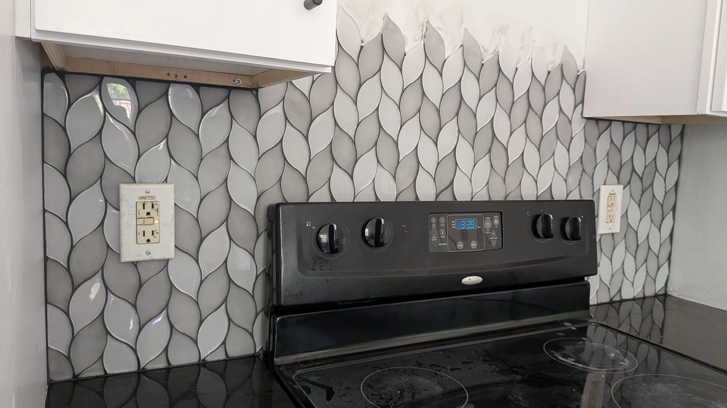 Kitchen countertop with a black electric stove, tiled backsplash with a leaf pattern in gray and white, electrical outlets on either side.