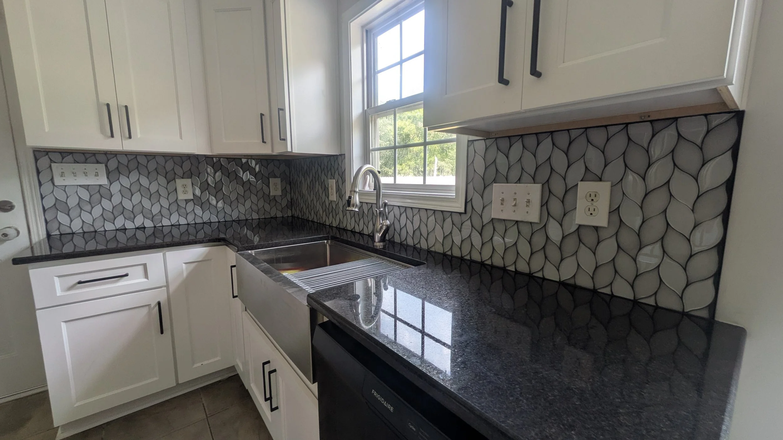 Kitchen with white cabinets, black and gray leaf-patterned backsplash, black granite countertop, stainless steel sink, and a window above the sink showing greenery outside.