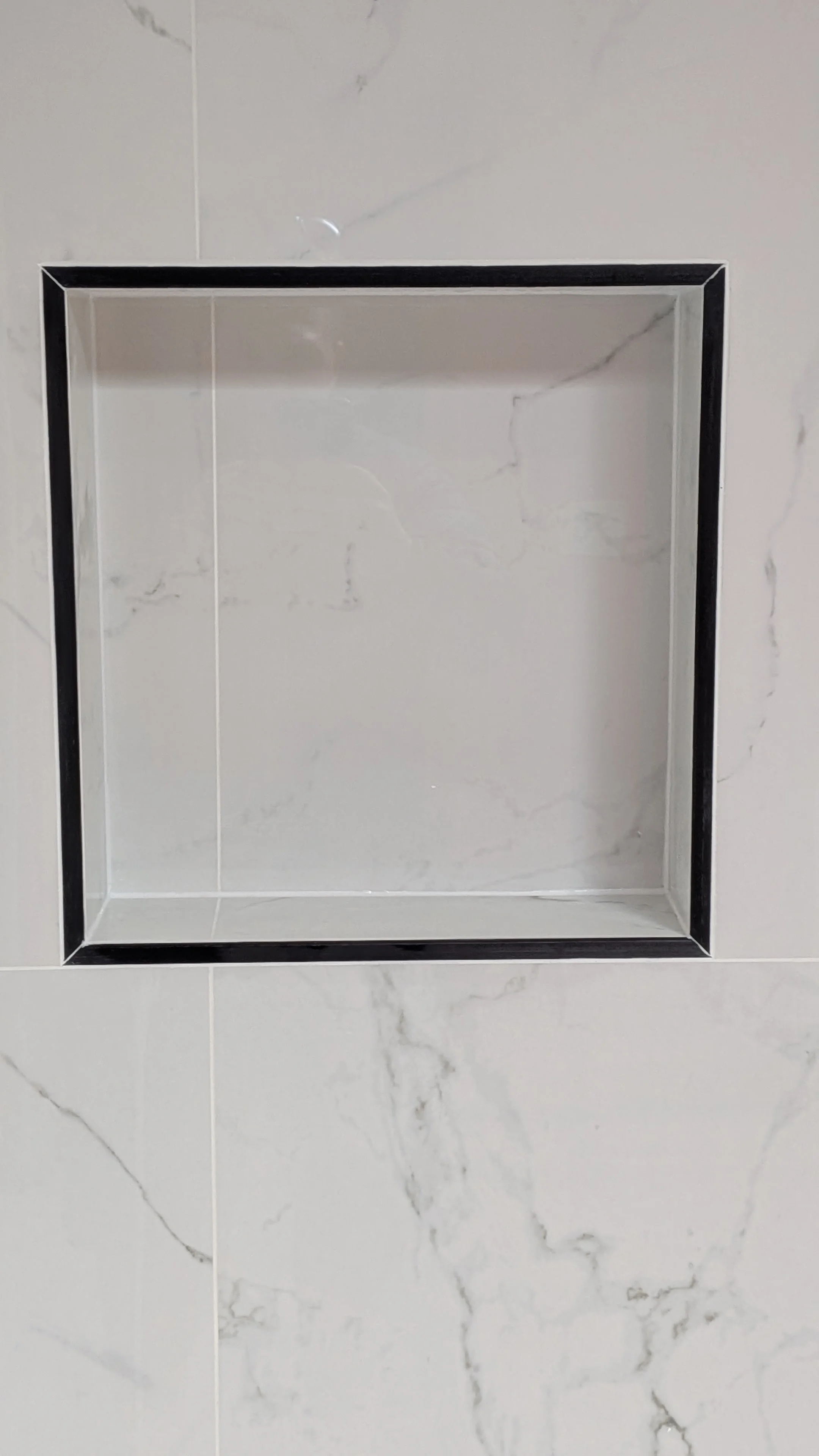 Empty rectangular shower niche with black trim on a white marble tiled wall.