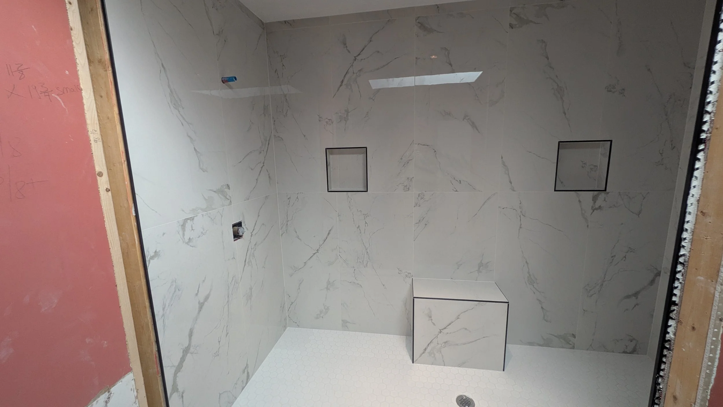 A shower with large white marble tiles on the walls, two built-in niches outlined in black, a small marble-topped bench, and white hexagonal floor tiles.