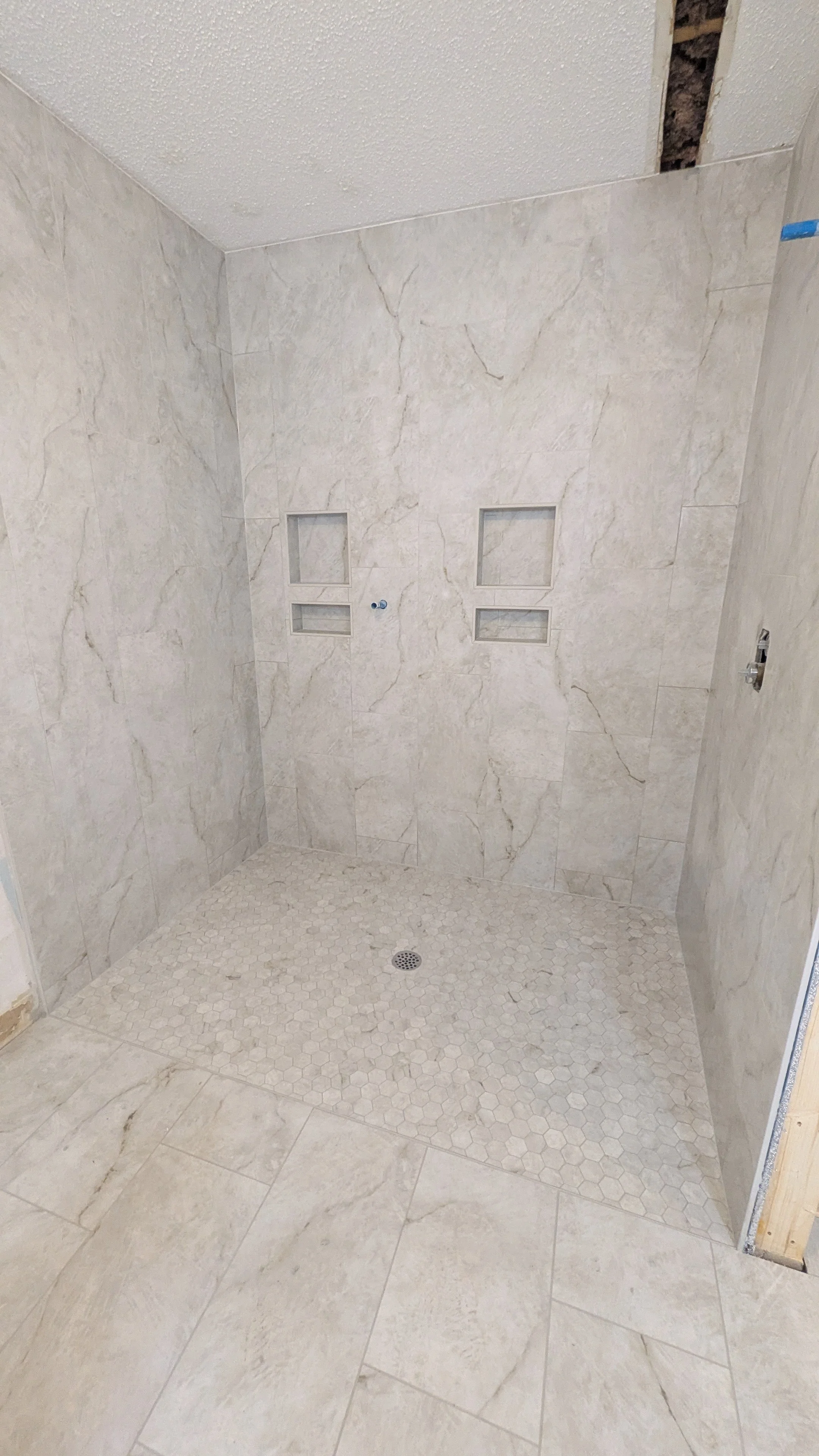 A newly tiled shower with beige marble tiles on the walls and white hexagonal tiles on the shower floor. Two built-in shelves are in the wall, with a pipe fitting and drain visible.