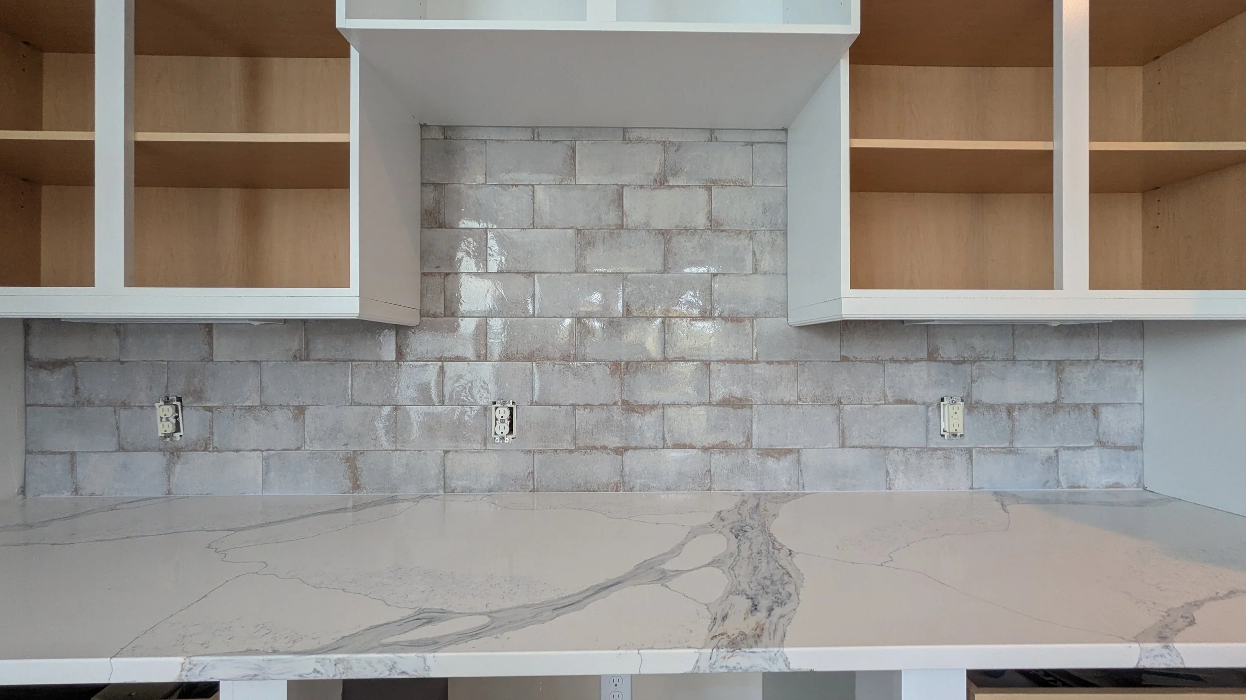 Kitchen backsplash with gray brick tiles, white upper cabinets without doors, electrical outlets, and a marble countertop with gray veining.
