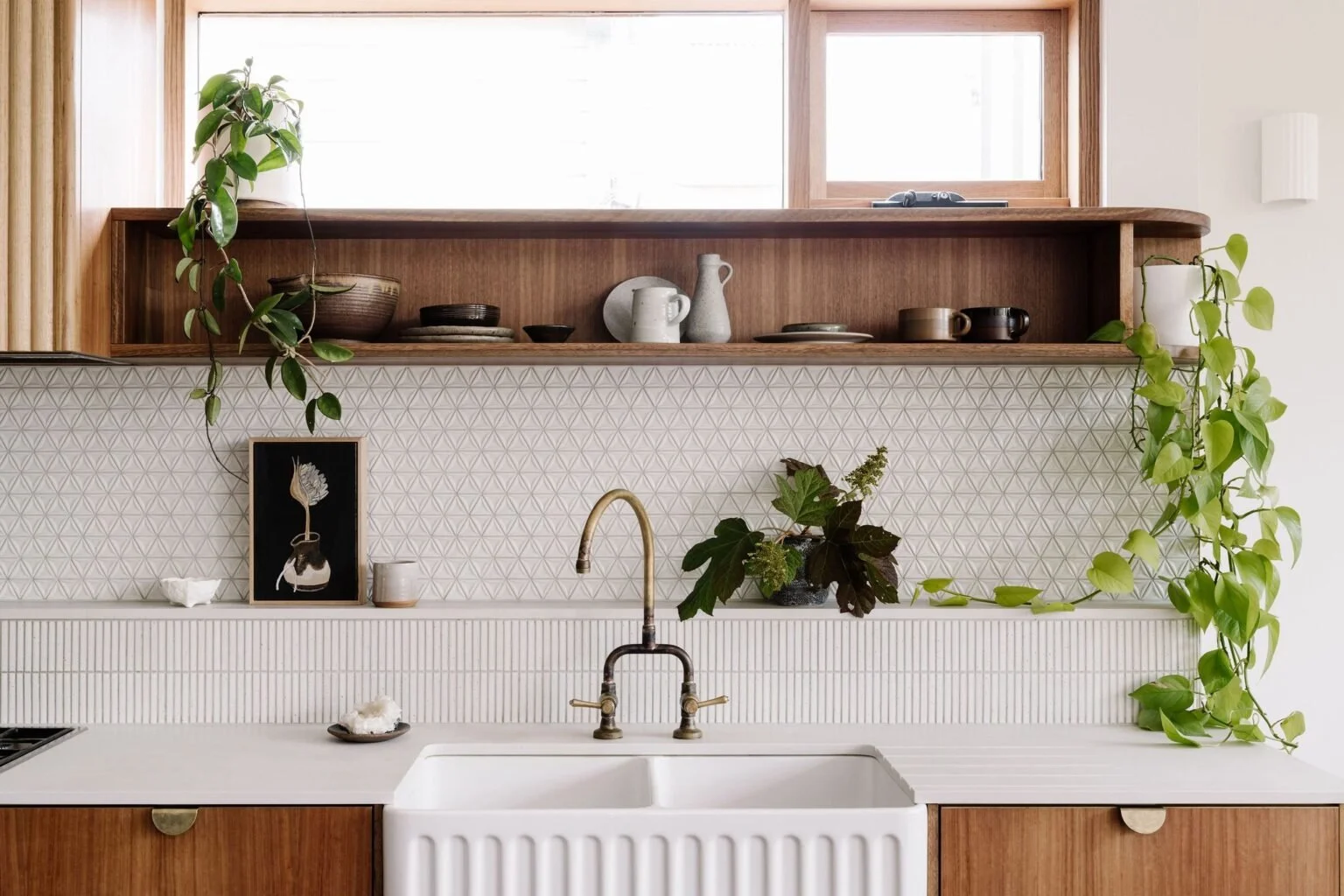 A modern kitchen with a white farmhouse sink, wooden cabinets, a brass faucet, and open wooden shelves holding various dishes and greenery, accented by geometric patterned tiles.