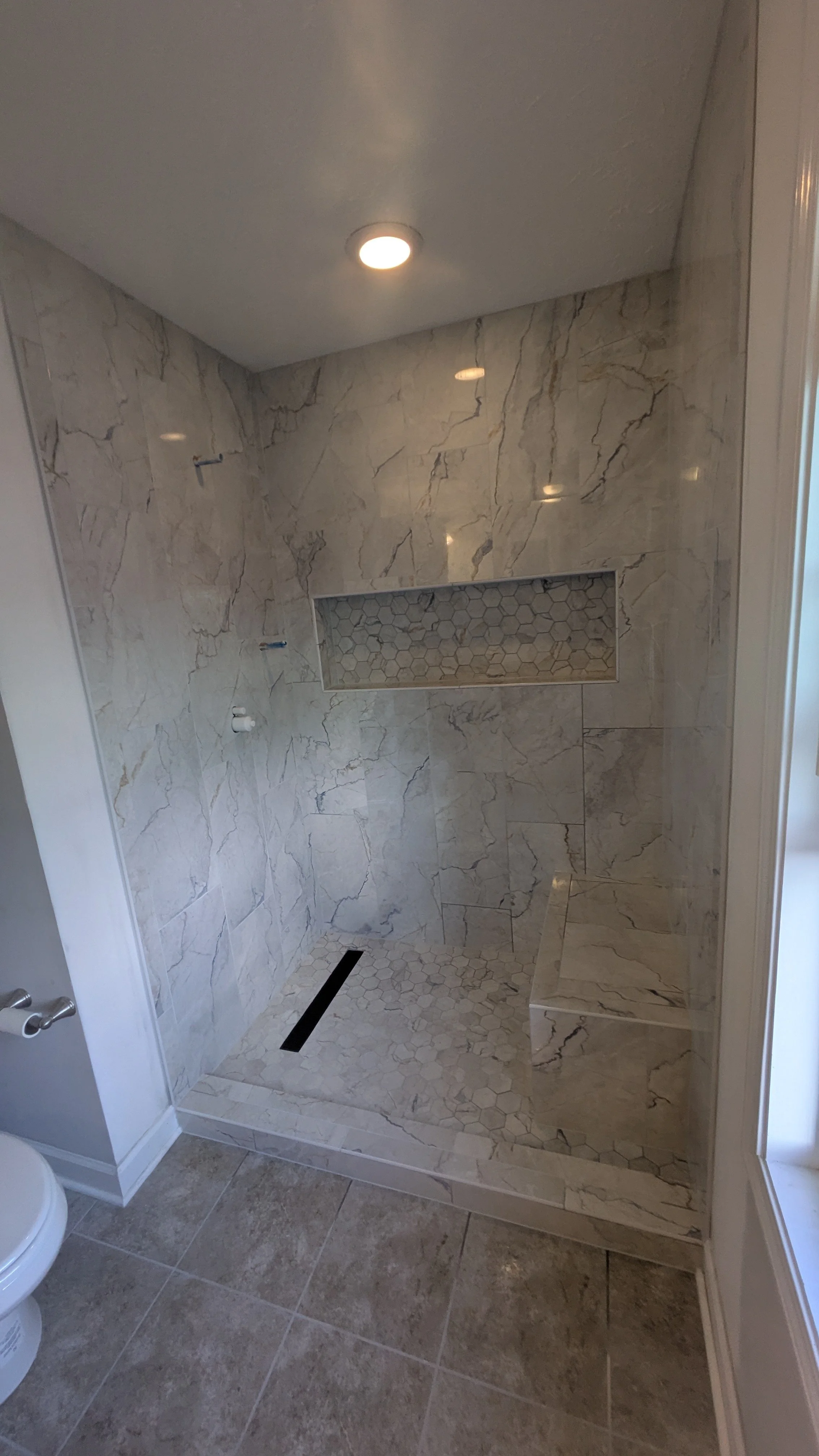 A modern walk-in shower with marble tiles on the walls and small hexagonal tiles on the floor and in the recessed niche. There is a drain in the floor and a bench to the right.