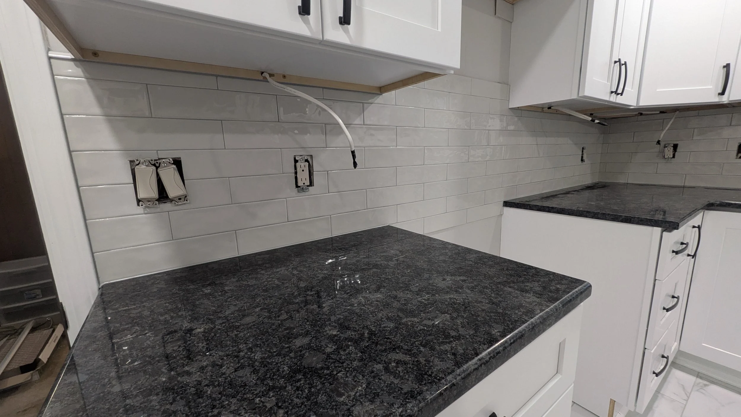 Kitchen with white cabinets, black granite countertops, white subway tile backsplash, and electrical outlets being installed.