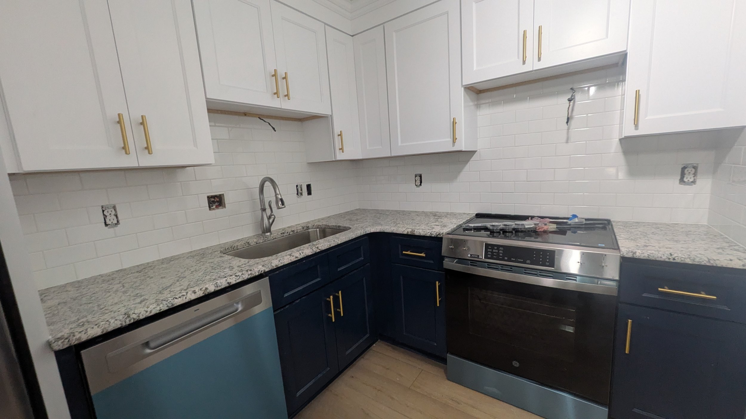 Kitchen with white upper cabinets, navy lower cabinets with gold handles, granite countertops, a stainless steel oven, and a small blue fridge.