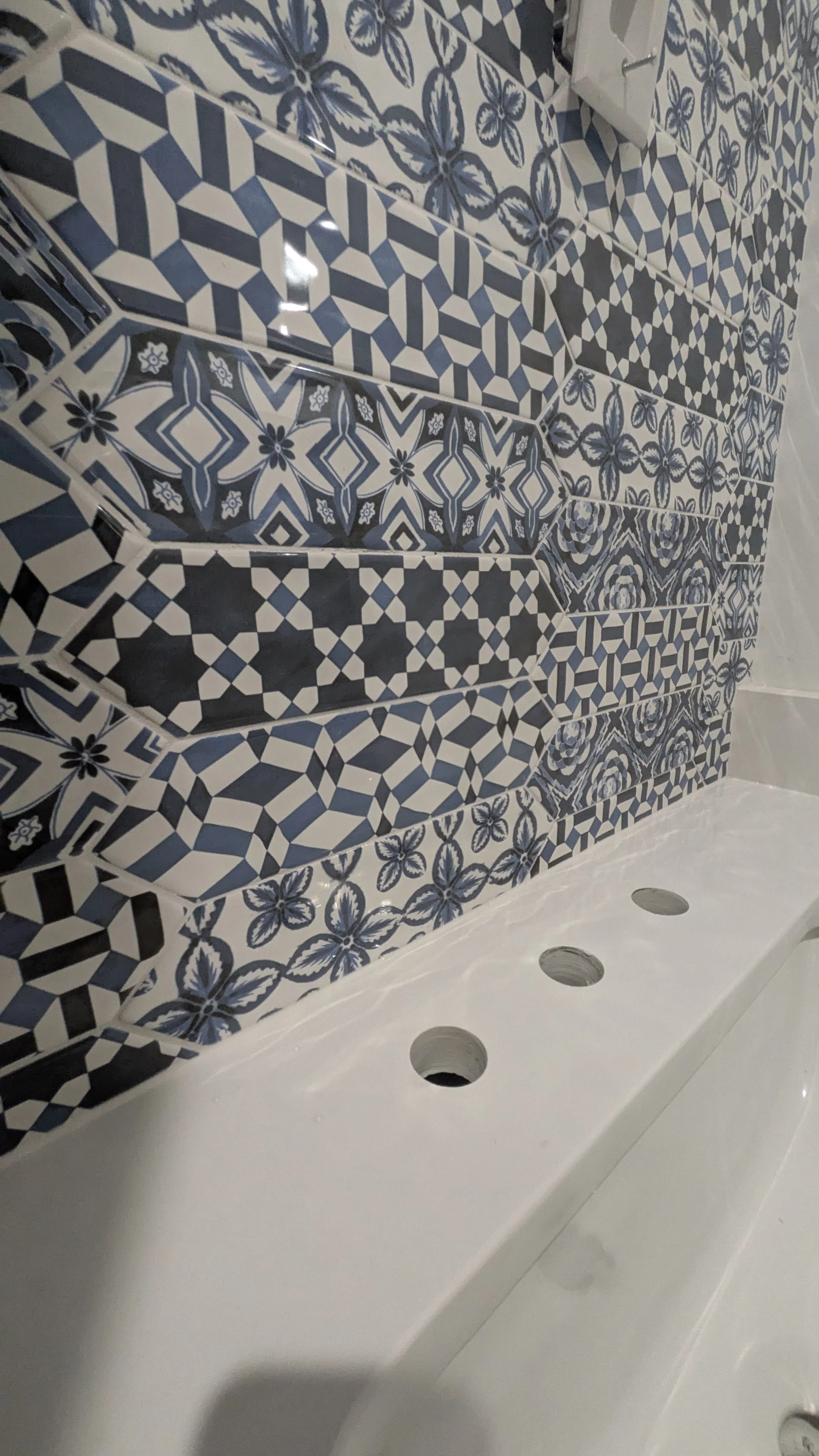 Close-up of a bathroom wall with decorative blue and white patterned tiles, featuring geometric and floral designs, above a white sink with three holes for faucets.