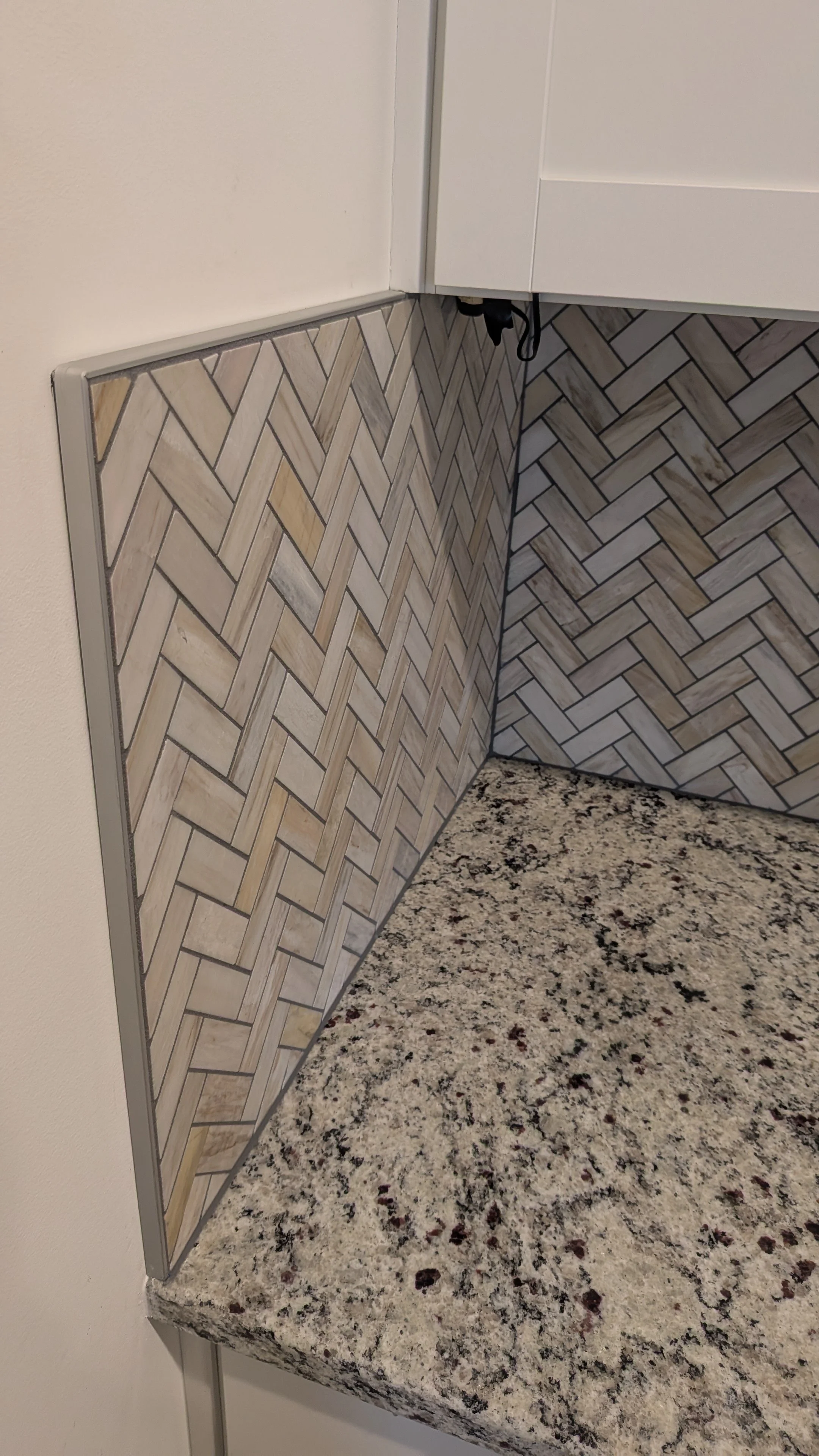 Close-up of a kitchen corner with a granite countertop, backsplash with wood-patterned tiles in a herringbone pattern, white cabinetry, and a wall outlet with cords hanging.