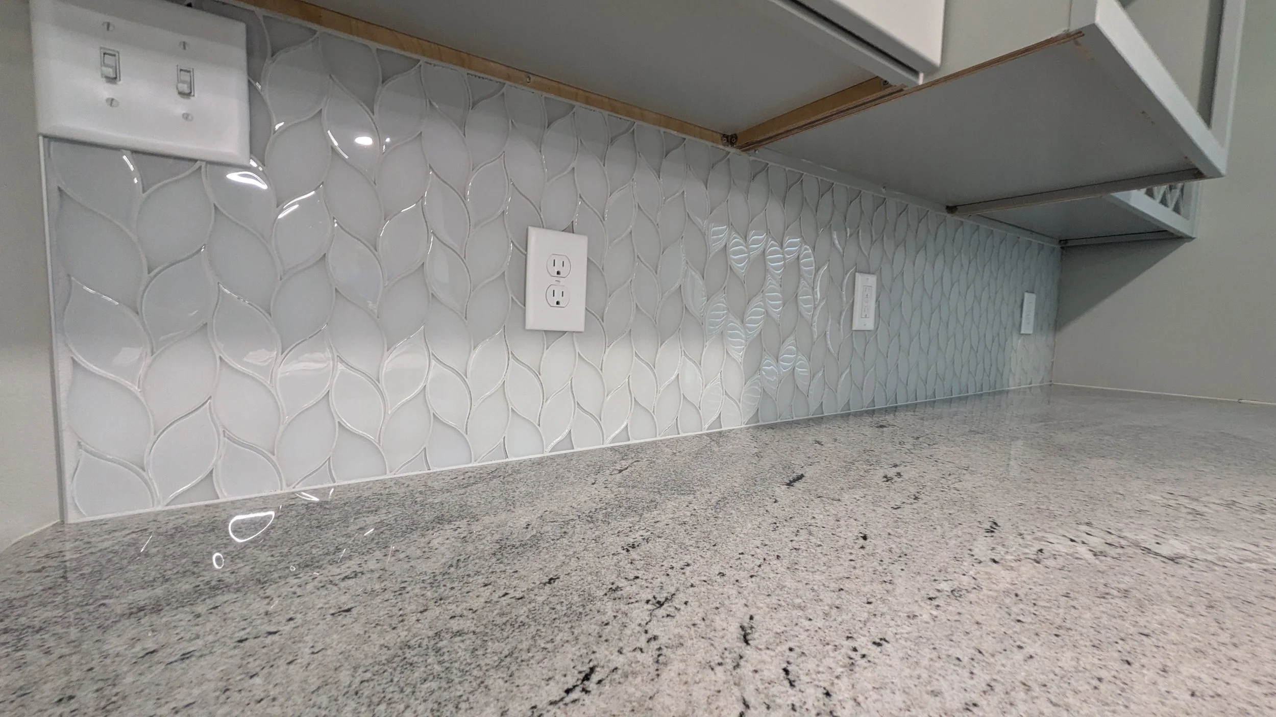 Close-up of a kitchen backsplash with white textured tiles and three electrical outlets, above a speckled granite countertop.