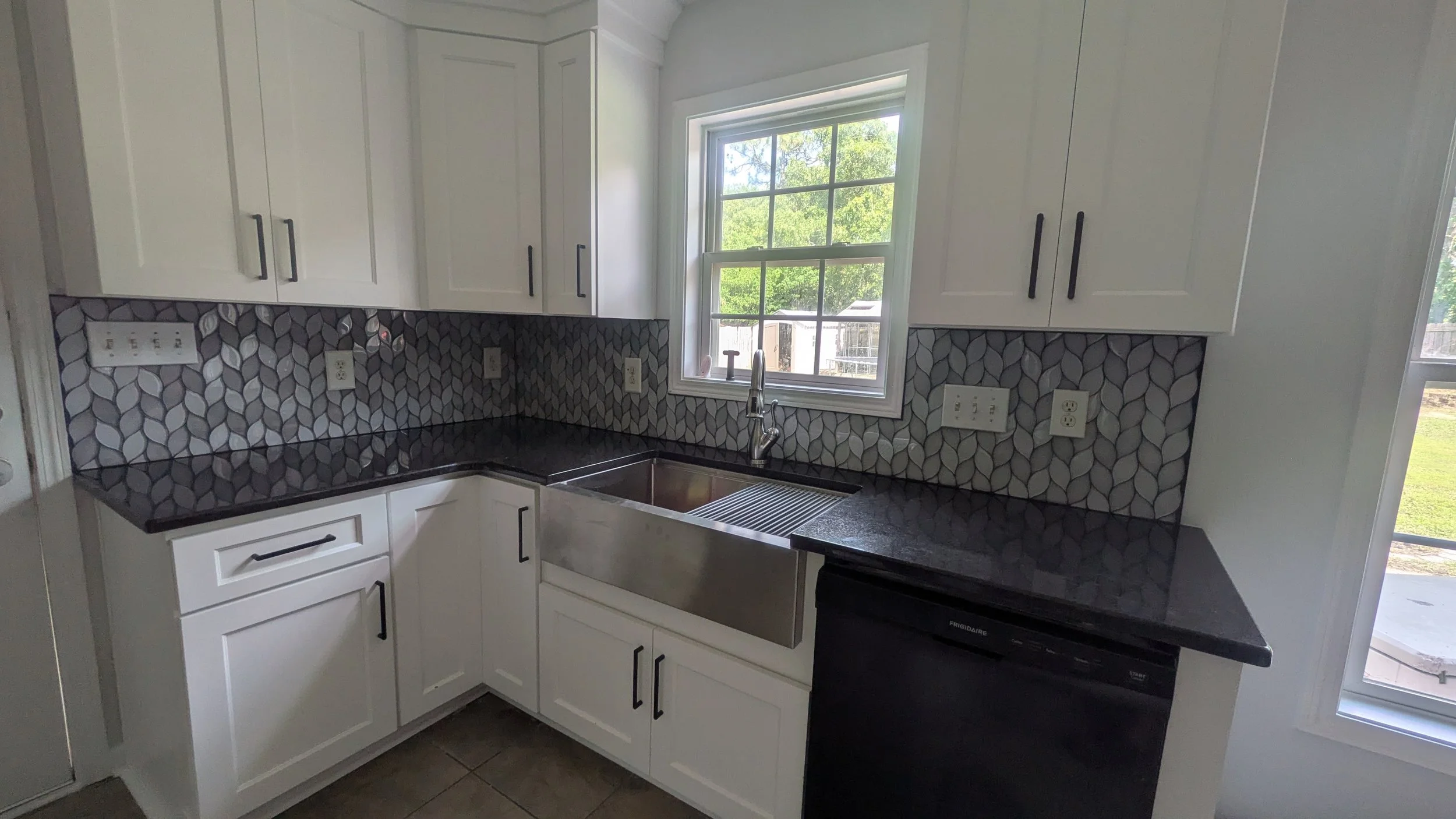 Kitchen with white cabinets, black countertop, stainless steel farmhouse sink, patterned backsplash, window above the sink, and multiple electrical outlets.