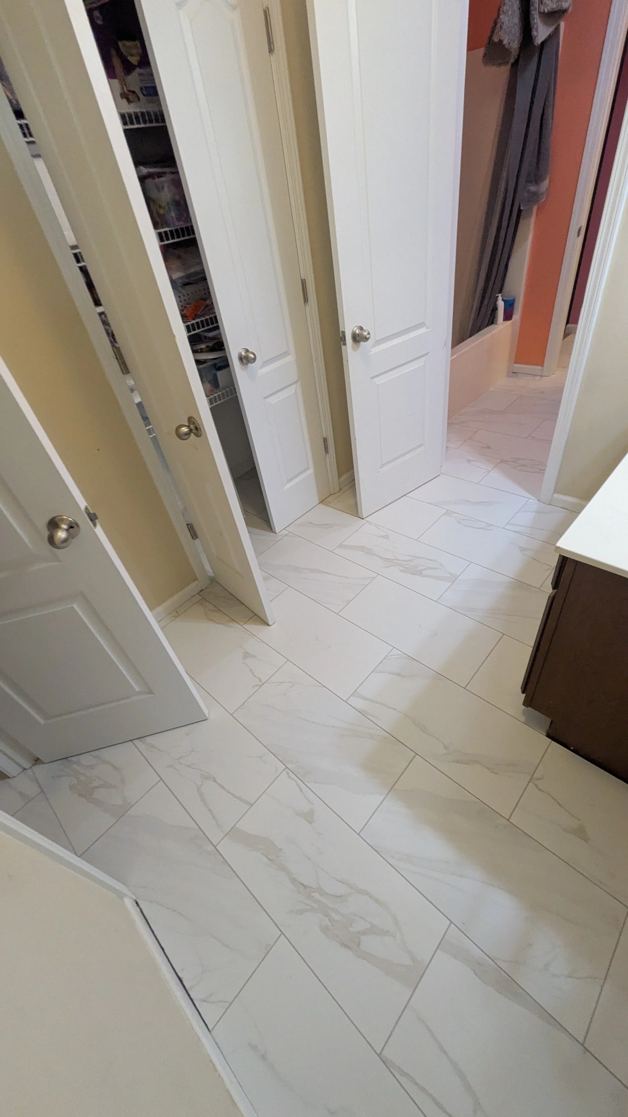 A bathroom with beige walls, white tiled floor with light gray veining, white cabinets, and an open closet door showing shelves with various items.